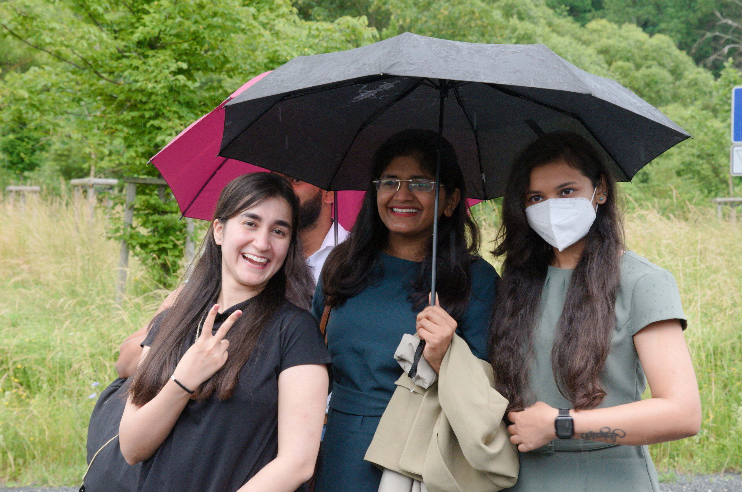 Drei Menschen stehen im Freien in der Nähe der Hochschule Coburg und lächeln unter Regenschirmen. Eine Person zeigt ein Peace-Zeichen, eine andere hält einen Regenschirm und die dritte trägt eine Gesichtsmaske. Sie sind an einem regnerischen Tag von Grün umgeben.