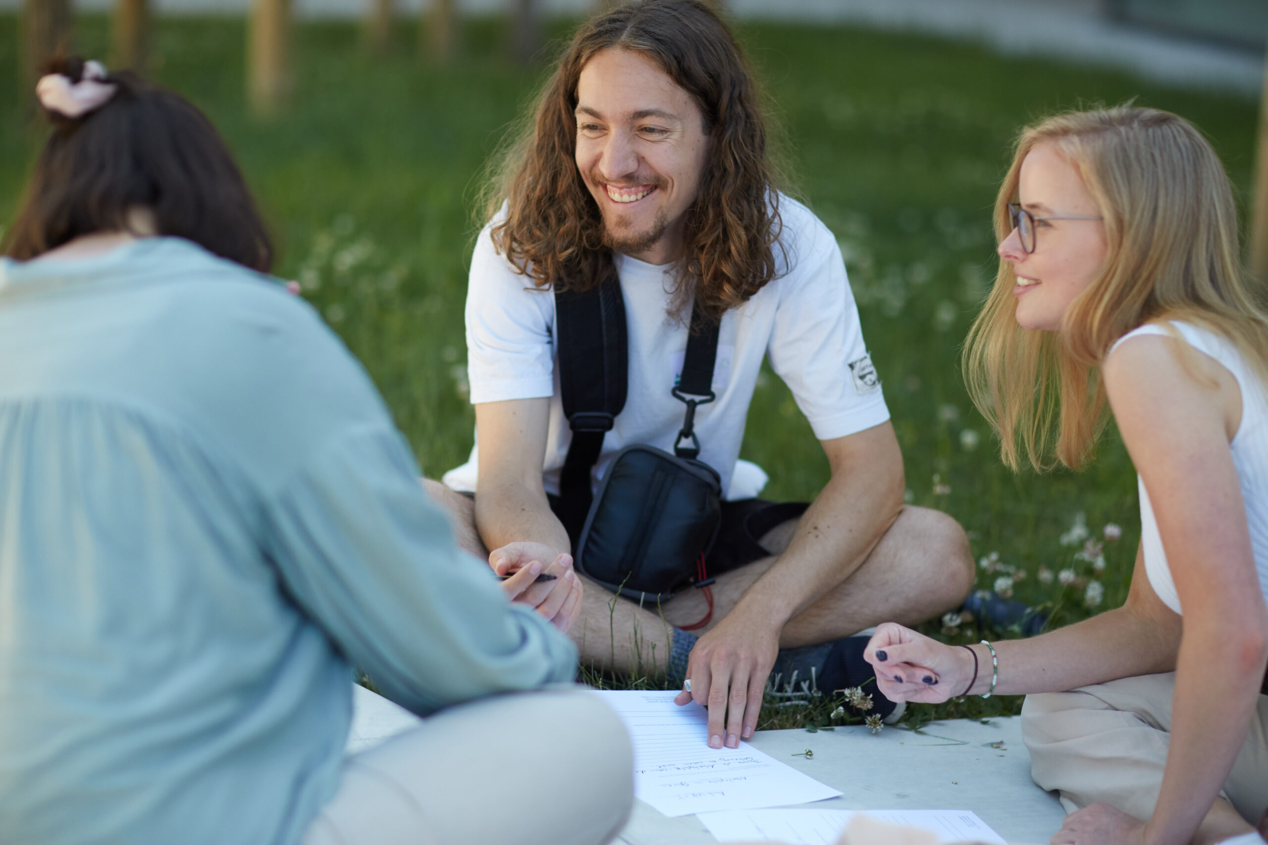 Drei Menschen sitzen auf der Wiese der Hochschule Coburg, unterhalten sich und schreiben auf Papier. Ein Mann mit langen Haaren lächelt, während zwei Frauen, eine davon mit Brille, aufmerksam zuhören. Sie befinden sich in einer entspannten Umgebung im Freien mit viel Grün im Hintergrund.