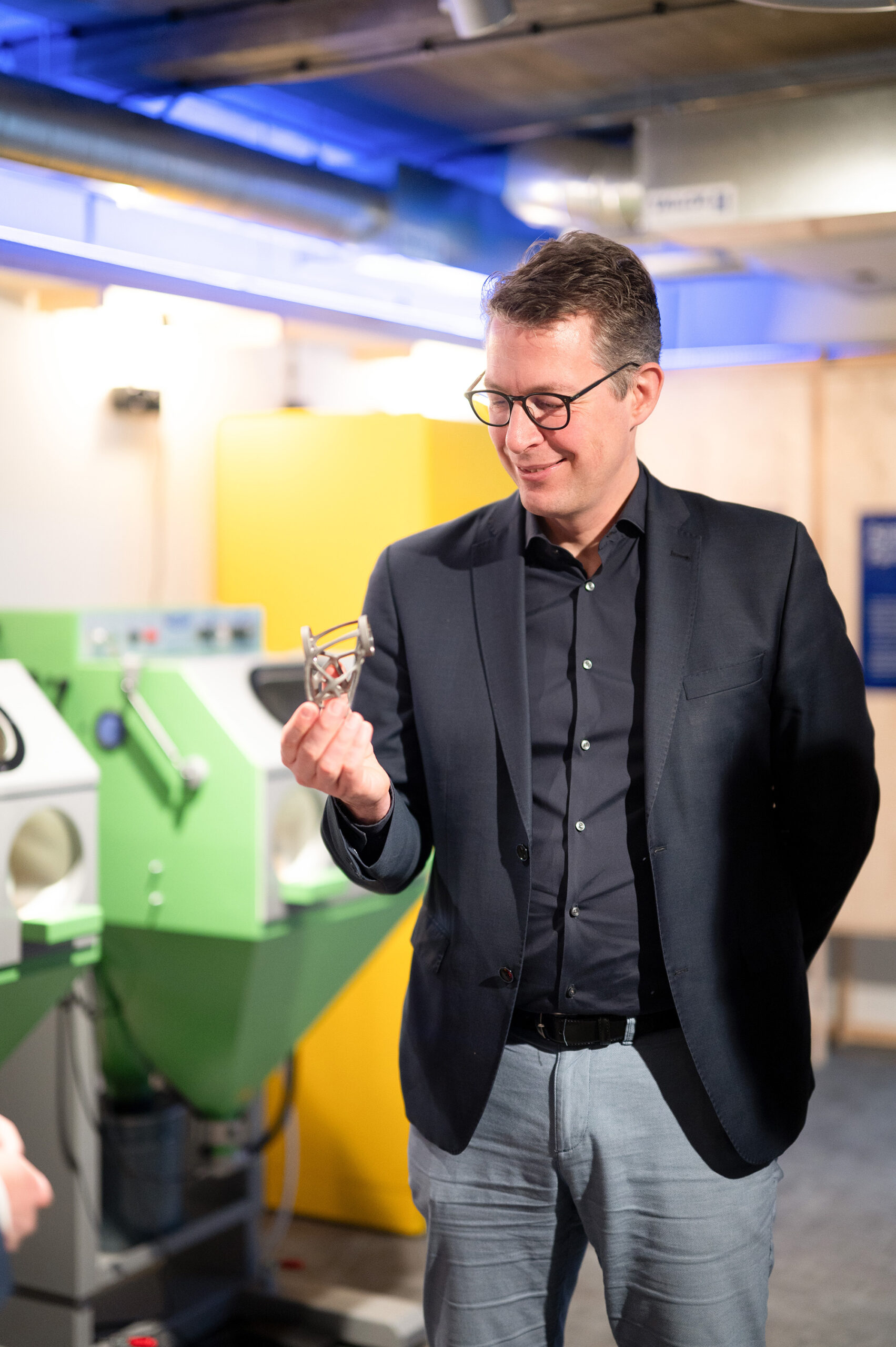 Ein Mann mit Brille und dunklem Anzug lächelt, während er in einem hell erleuchteten Labor der Hochschule Coburg einen kleinen metallischen Gegenstand hält. Im Hintergrund sind grün-gelbe Maschinen zu sehen, die die hochmoderne Innovation hervorheben, die dort stattfindet.