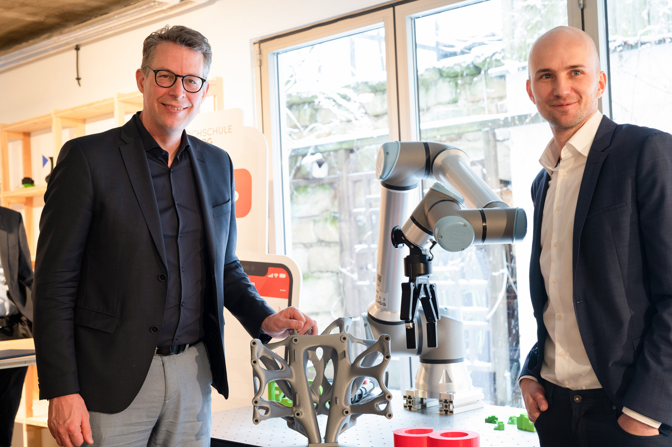 Two men in suits stand next to a robotic arm holding metal parts in the modern, well-lit office of Hochschule Coburg, with large windows. The table is scattered with various mechanical components and small green objects.