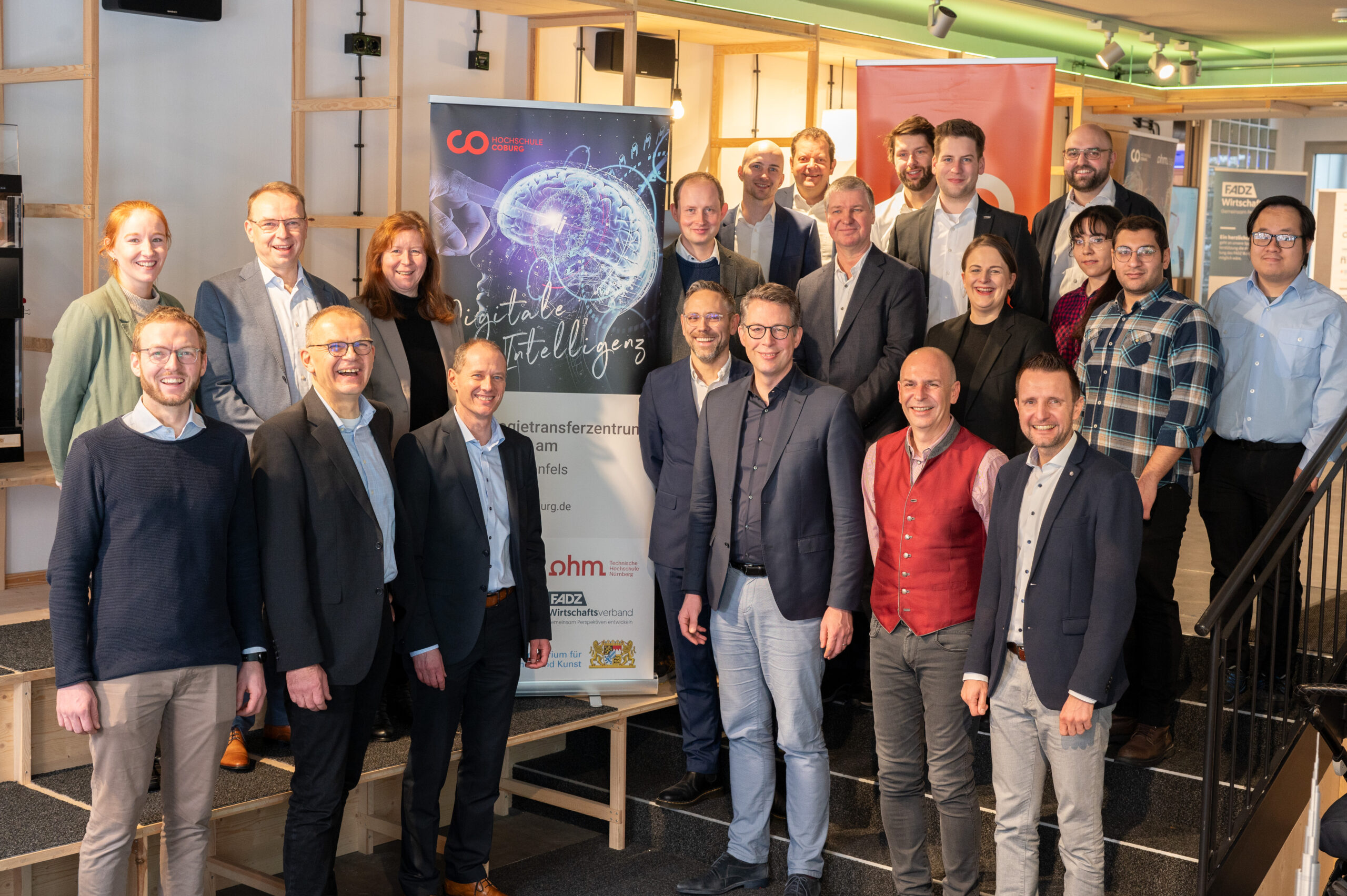 A group of people, both men and women, pose together indoors at Hochschule Coburg. Behind them is a large poster with an image of a brain and the words "Künstliche Intelligenz," alongside a "CO" logo. The setting appears to be an office or conference room.