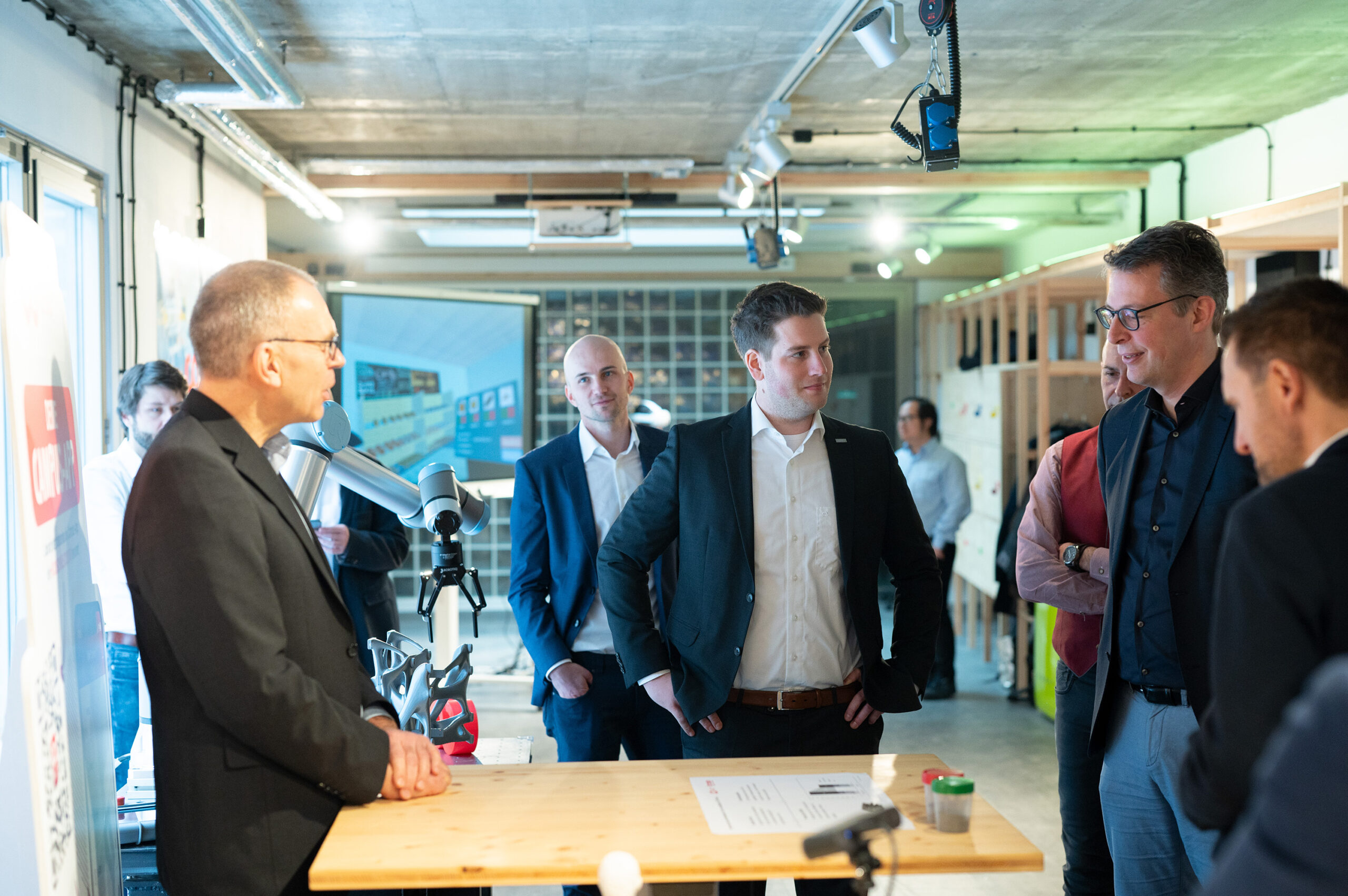 A group of professionally dressed individuals engage in conversation around a wooden table in a modern office at Hochschule Coburg. A robot arm and a presentation screen are visible in the background, highlighting the innovative environment.