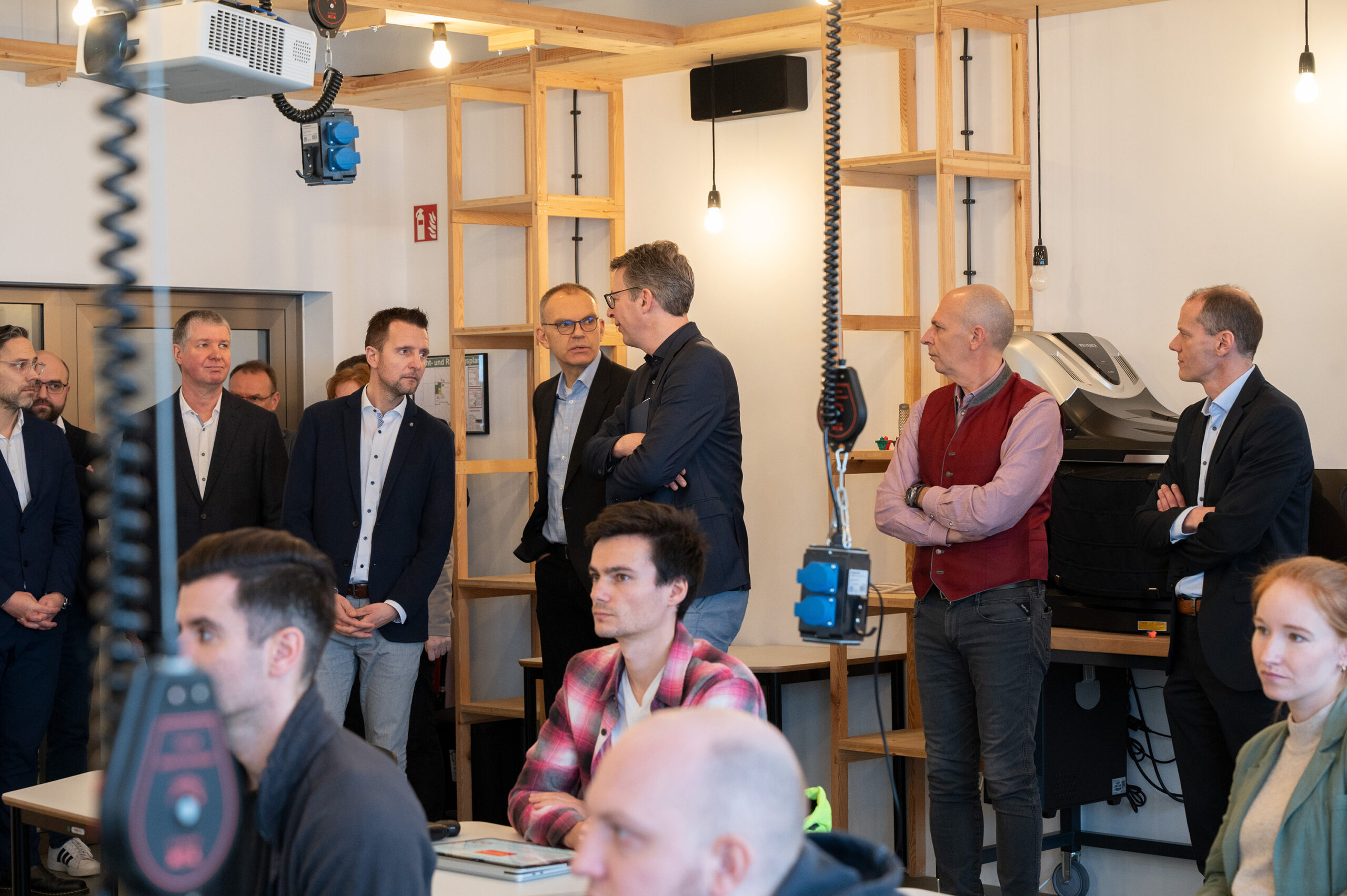 A group of people in business attire are standing and conversing in a modern office space at Hochschule Coburg. Others are seated, listening attentively. The room features wooden shelving, exposed bulbs, and technical equipment.