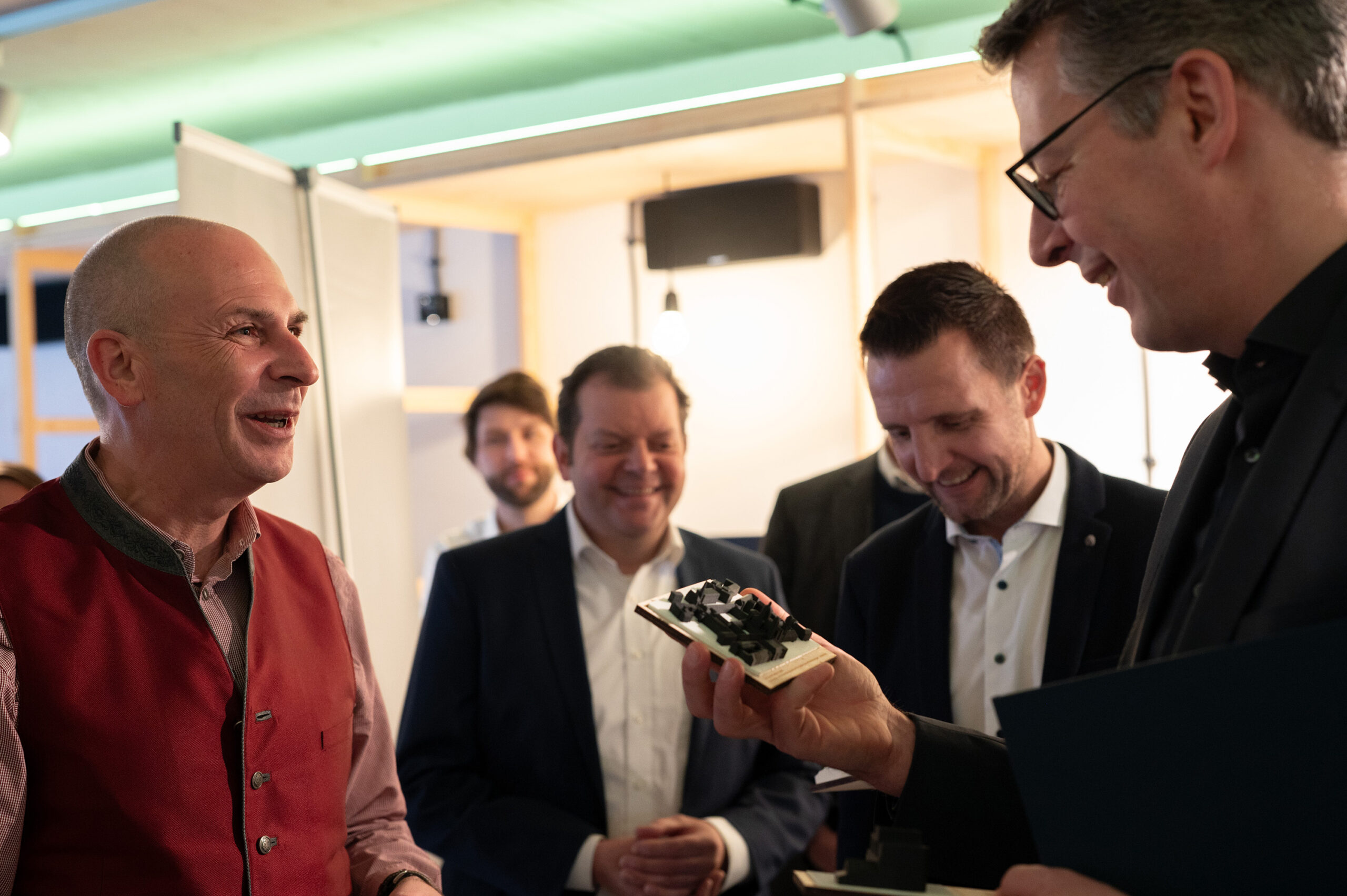 A group of five men in suits and casual wear are smiling and engaged in conversation at Hochschule Coburg. One man holds an item resembling a small sculpture. They appear to be in a modern indoor setting with soft lighting, creating an atmosphere of intellectual exchange.