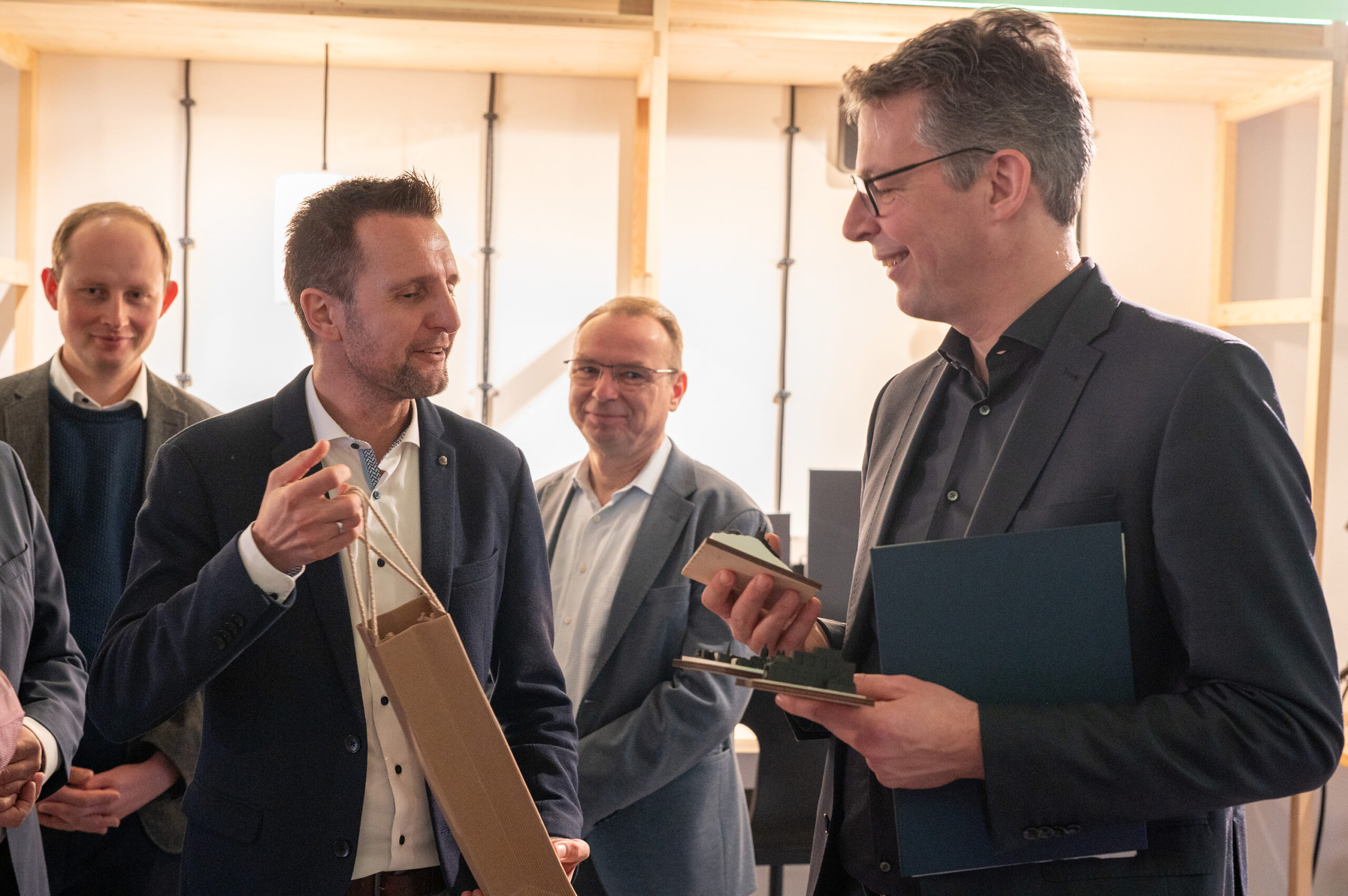 A group of five men in business attire stand together in an office setting at Hochschule Coburg. Two of them are interacting, one holding a gift bag and the other a notebook and folder. They appear to be engaged in a pleasant conversation.