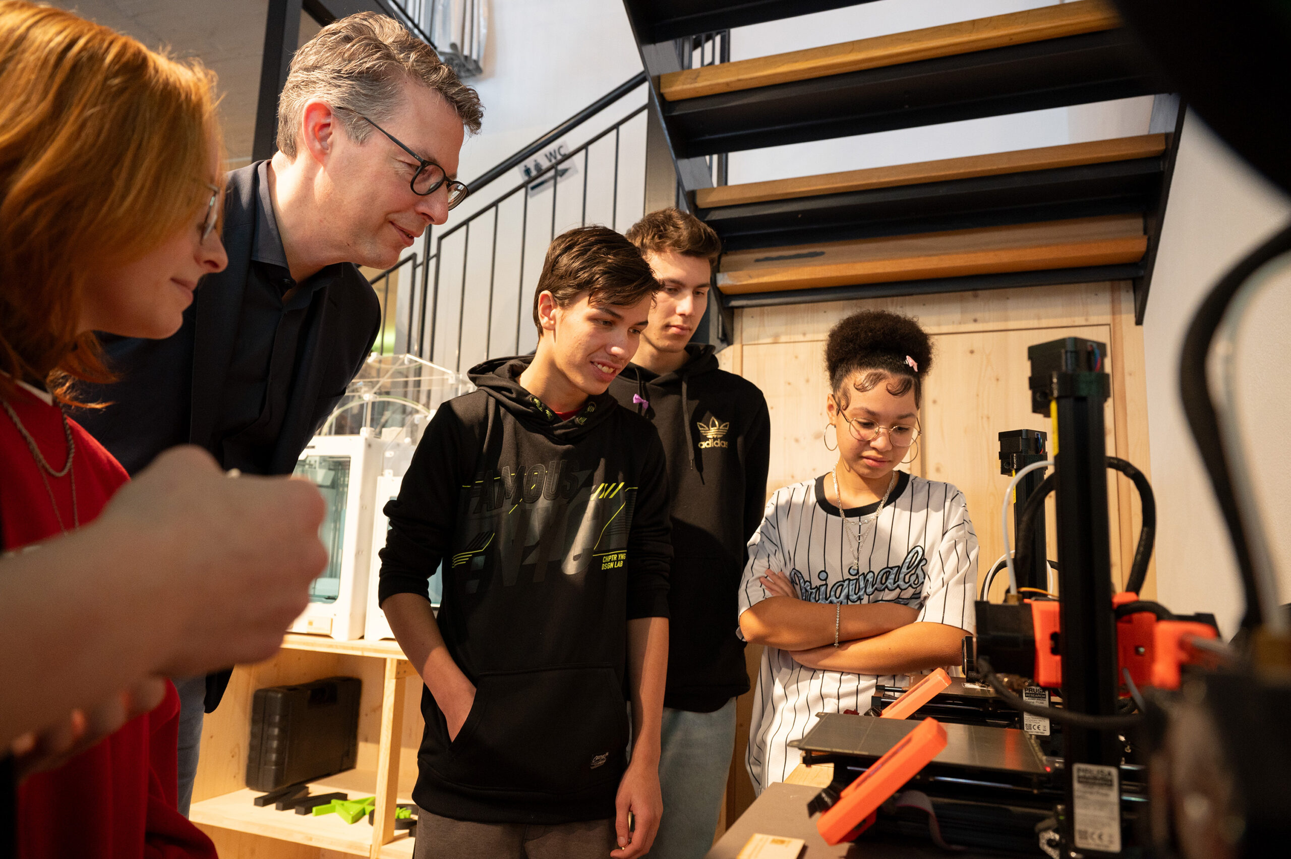 Eine Gruppe von Studierenden versammelt sich mit einem Dozenten der Hochschule Coburg um einen 3D-Drucker. In der Werkstattatmosphäre beobachten sie interessiert den Druckvorgang, während Regale und eine Treppe eine dynamische Kulisse schaffen.