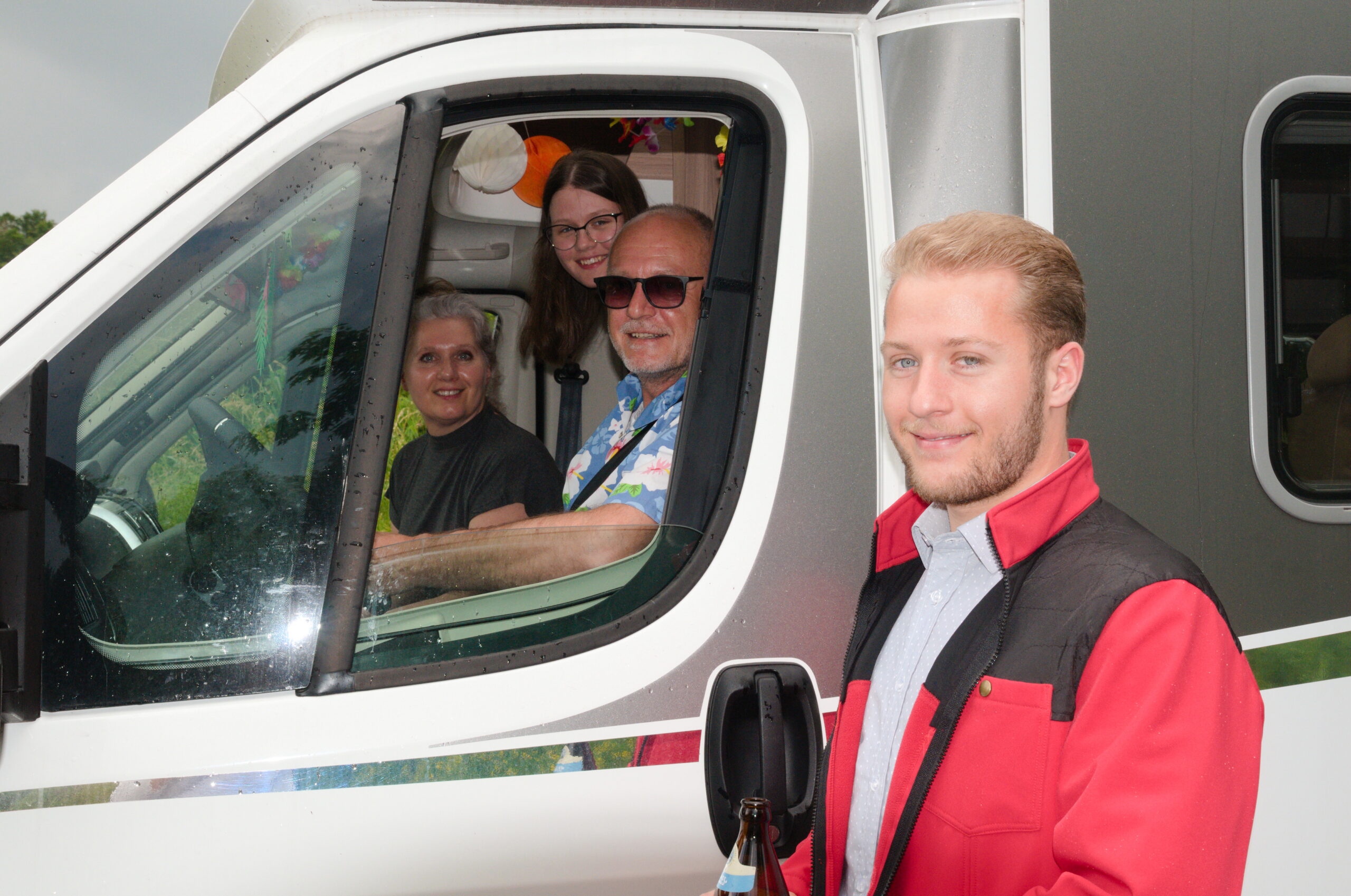 Ein lächelnder junger Mann in einer roten Jacke steht vor einem Wohnmobil in der Nähe der Hochschule Coburg. Im Inneren lächeln drei Menschen in die Kamera: ein Mann mit Sonnenbrille, eine Frau mit kurzen grauen Haaren und eine Frau mit langen braunen Haaren. Der Innenraum ist mit farbenfrohen Gegenständen dekoriert.