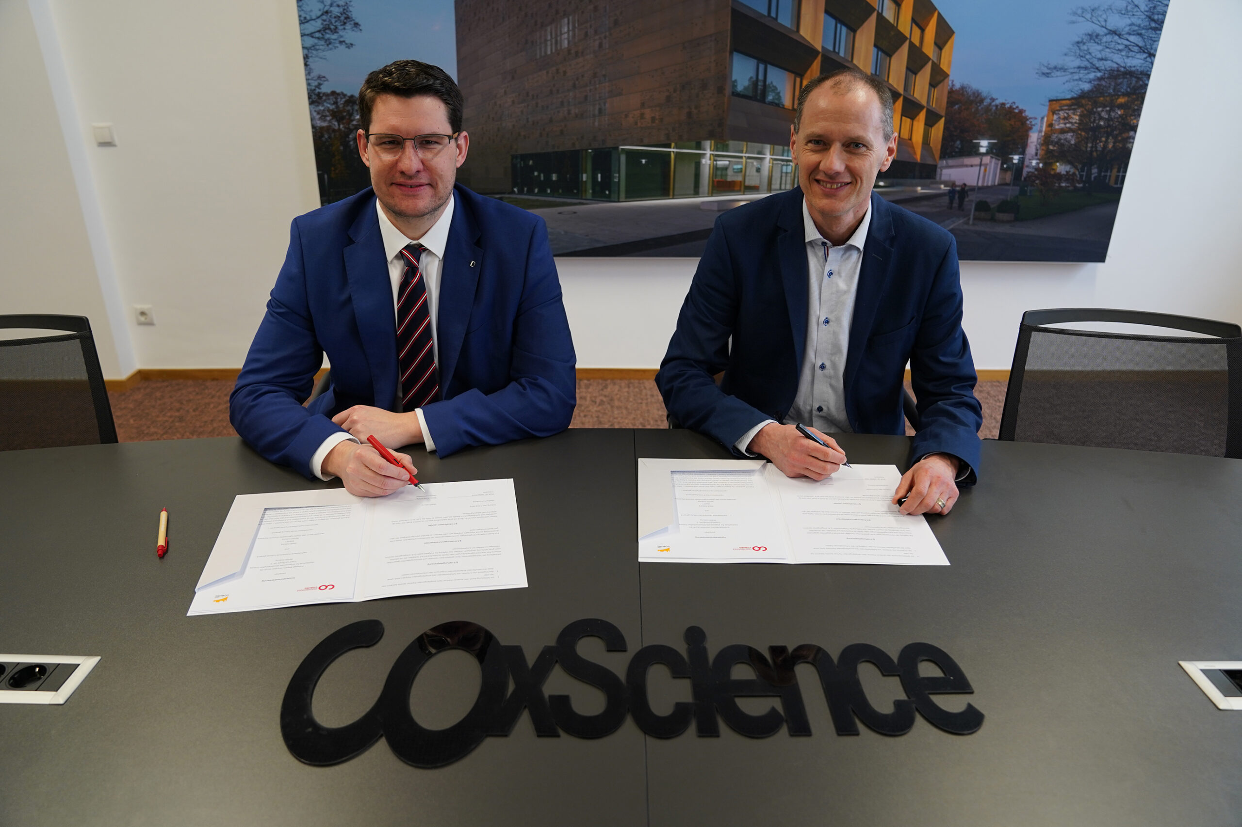 Two men in suits are seated at a table, signing documents. The table features the text "coXscience." Behind them, a large photo of Hochschule Coburg's iconic building is prominently displayed on the wall.