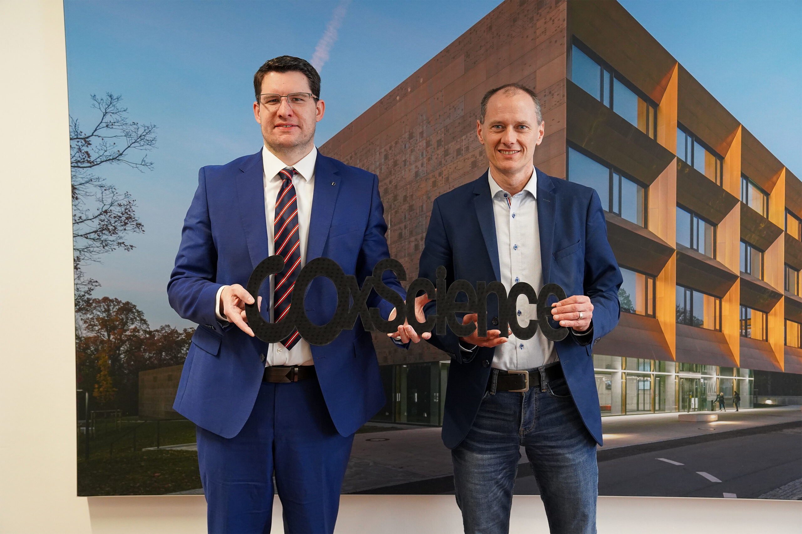 Two men in suits stand indoors holding a sign that reads "GoScience," set against a backdrop showcasing the modern architecture of Hochschule Coburg.