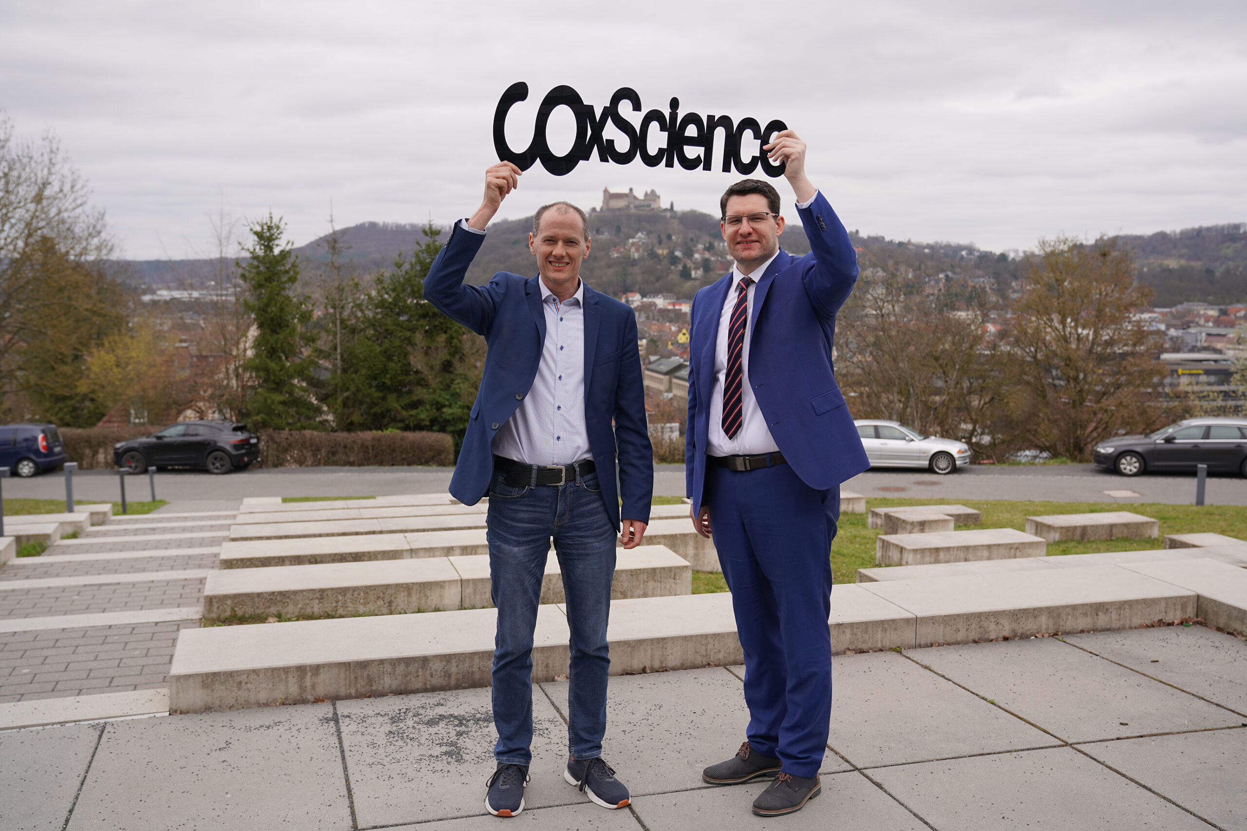 Two men stand outdoors, both wearing blue suits. One holds a cutout sign above their heads reading "CoScience." They are on a paved area at Hochschule Coburg with a scenic landscape in the background, featuring hills and buildings under a cloudy sky.