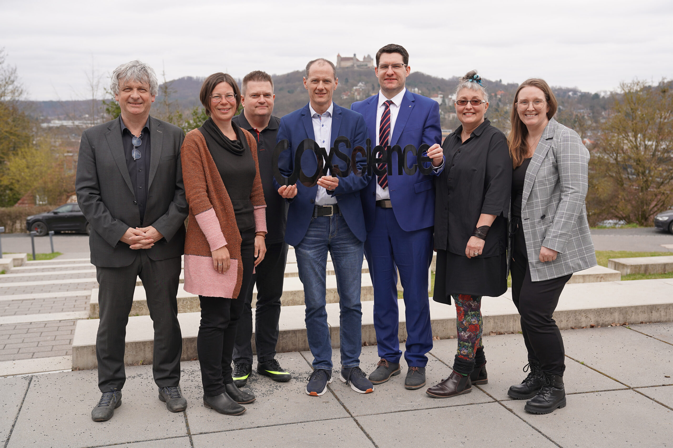 A group of seven people stands together outdoors on a paved area, smiling and holding the word "Science" in cut-out letters. Behind them, a scenic view reveals a hill topped by a castle under a cloudy sky, reminiscent of those near Hochschule Coburg.