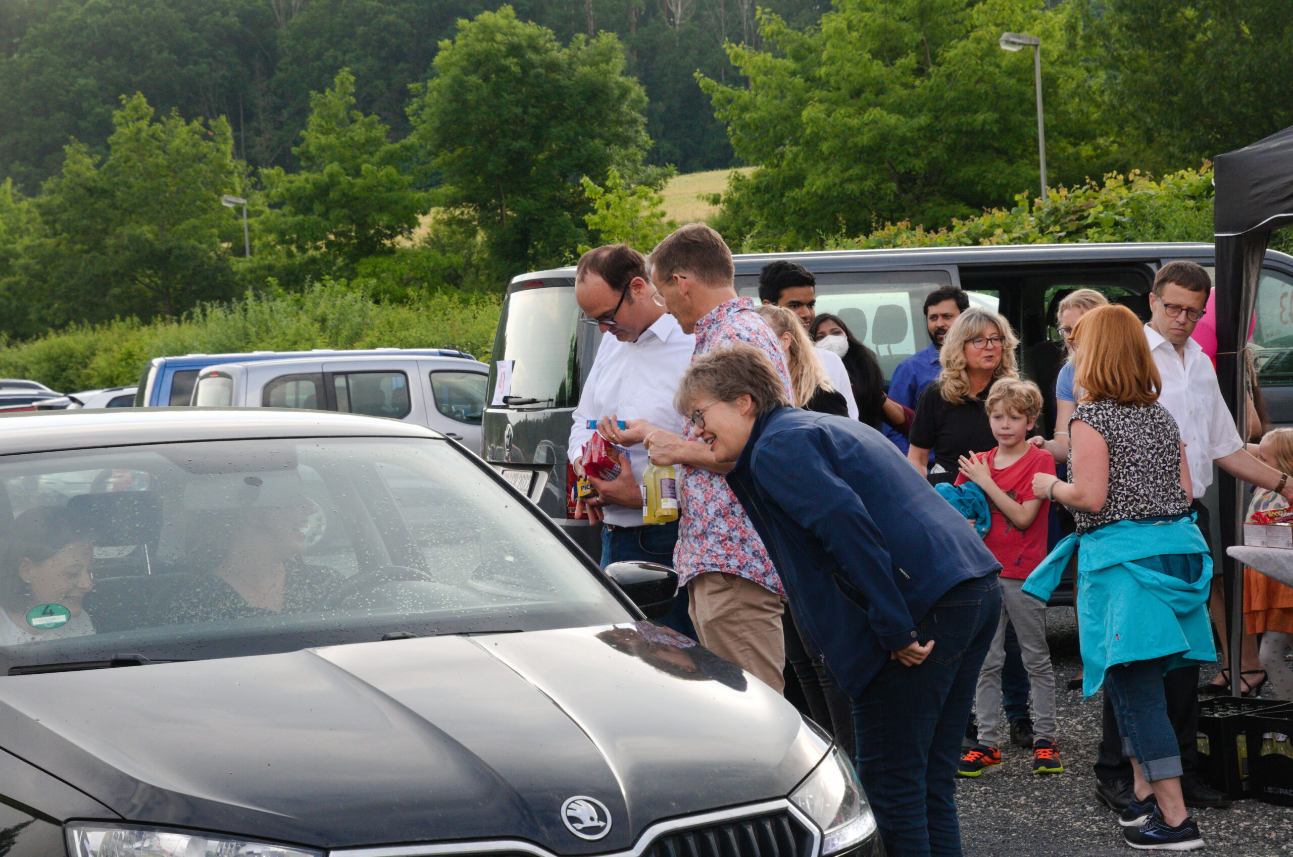 Eine Gruppe von Menschen versammelt sich um ein schwarzes Auto, das auf dem Parkplatz der Hochschule Coburg geparkt ist. Einige unterhalten sich mit den Menschen im Auto, während Bäume und andere Fahrzeuge eine malerische Kulisse bilden.