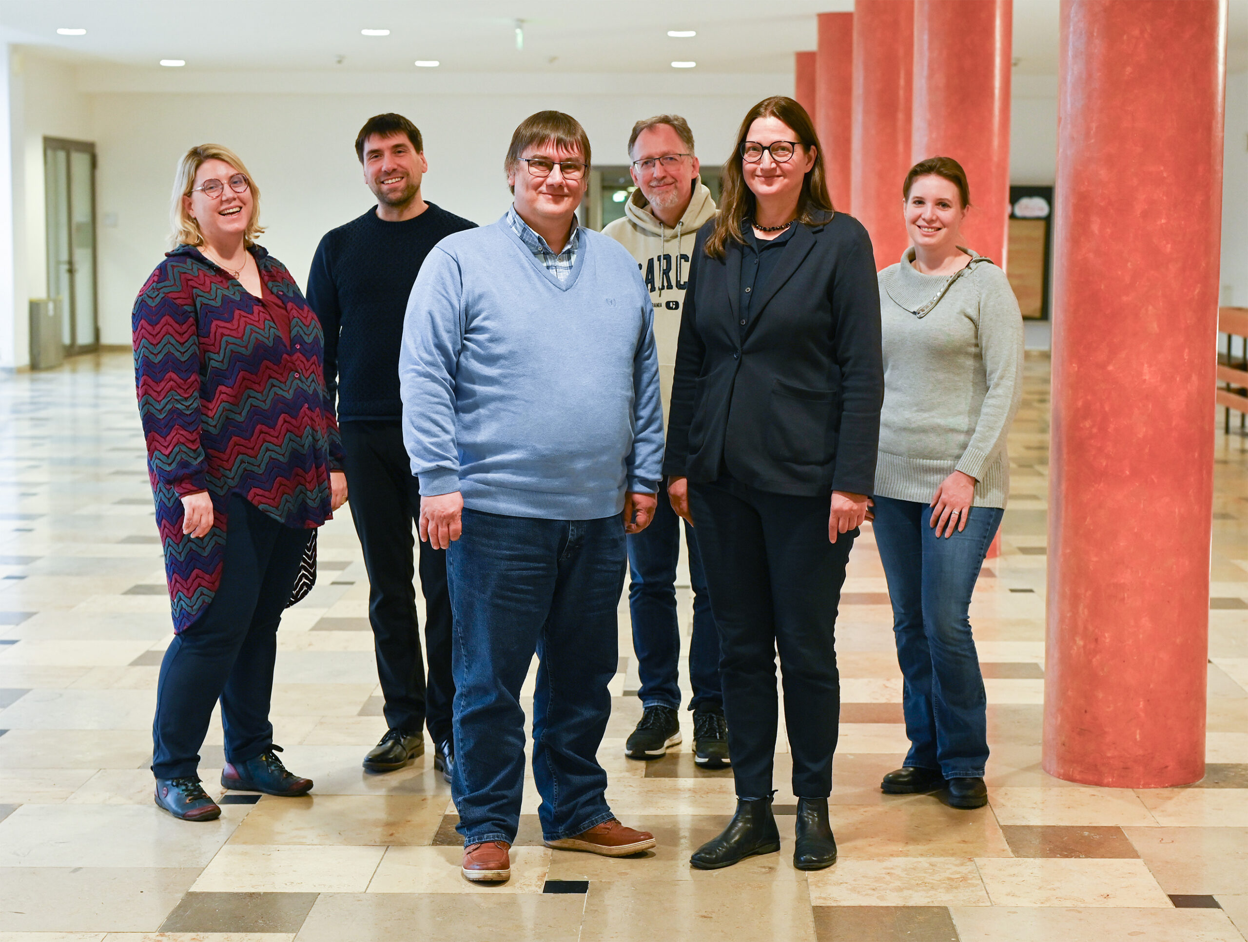 A group of six people stands in a bright hallway at Hochschule Coburg, with red columns in the background. They are smiling and casually dressed, enjoying the academic setting. The floor is tiled in a light color.