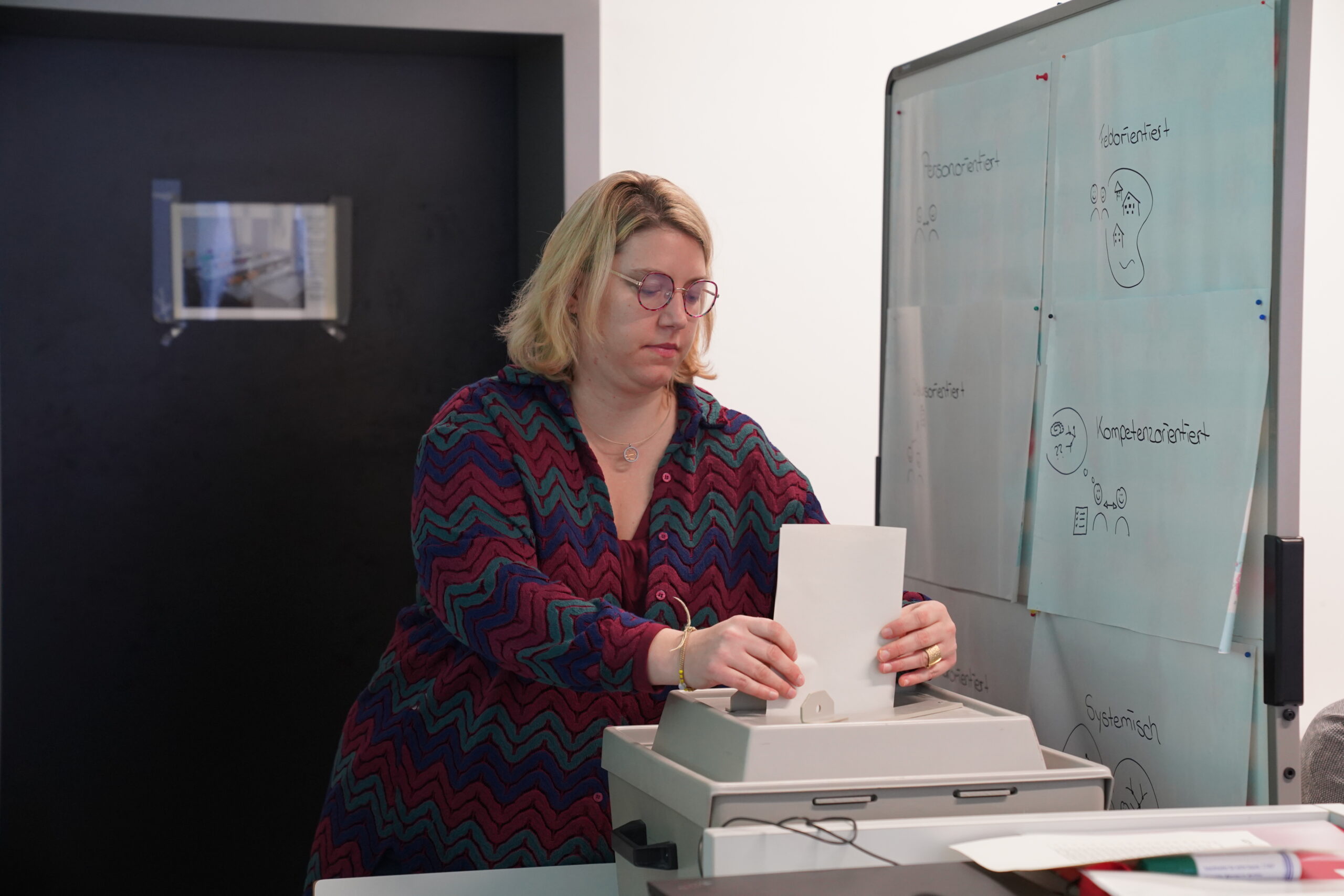 A person with shoulder-length blonde hair and glasses operates a paper shredder in an office at Hochschule Coburg. Nearby, a whiteboard displays various drawings and notes. A small stack of papers and other office supplies are visible on the table.