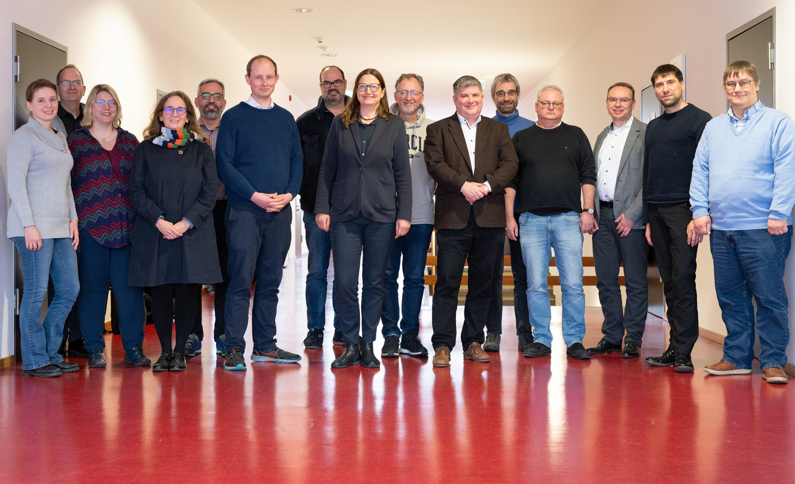 A group of 15 people, both men and women, stand in the brightly lit hallway of Hochschule Coburg, which features a striking red floor. Casually dressed, they face the camera with expressions that are friendly and relaxed.