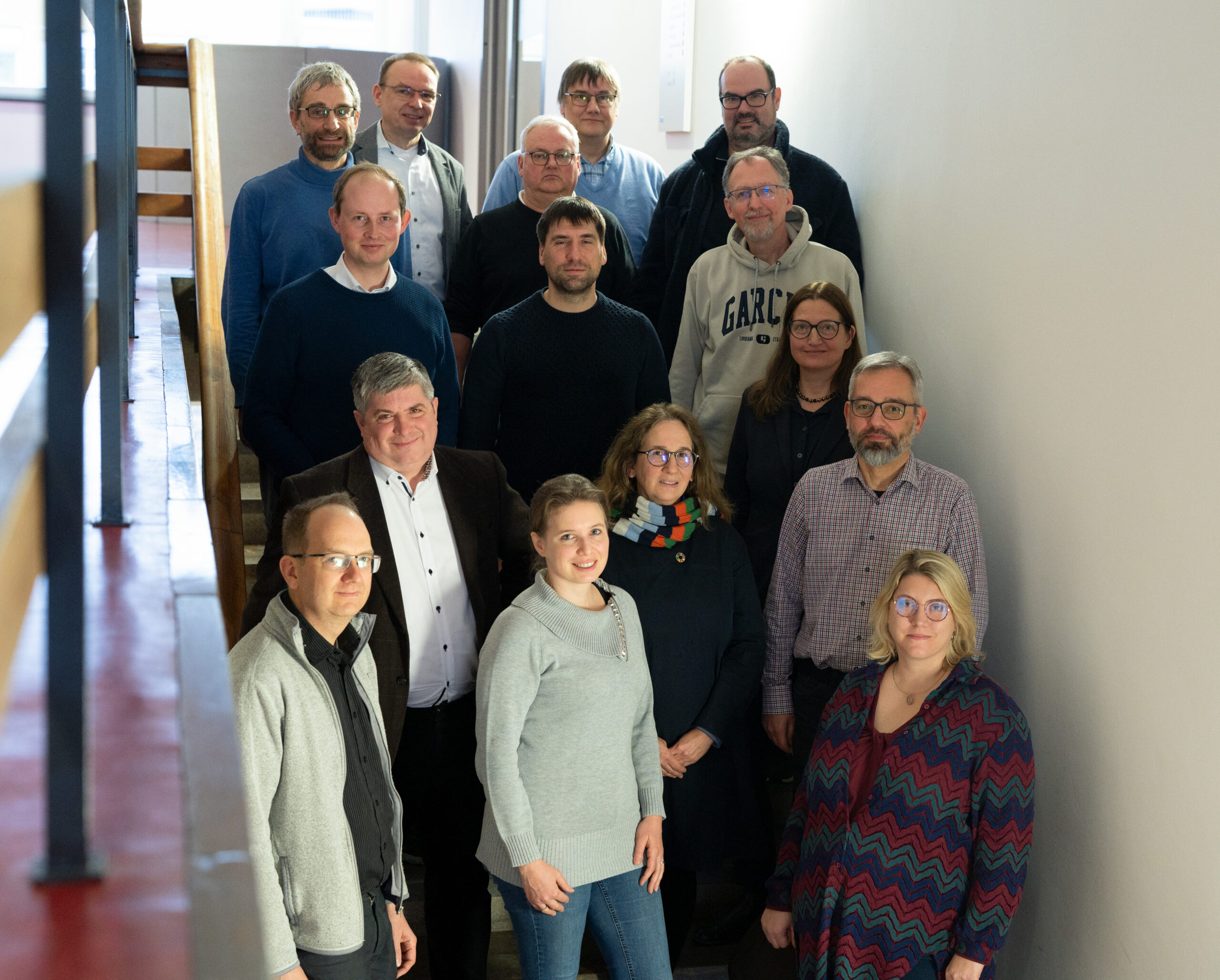 A group of thirteen people, standing in two rows on a staircase at Hochschule Coburg, pose for a photo indoors. They are dressed in casual clothing, and most are wearing glasses.