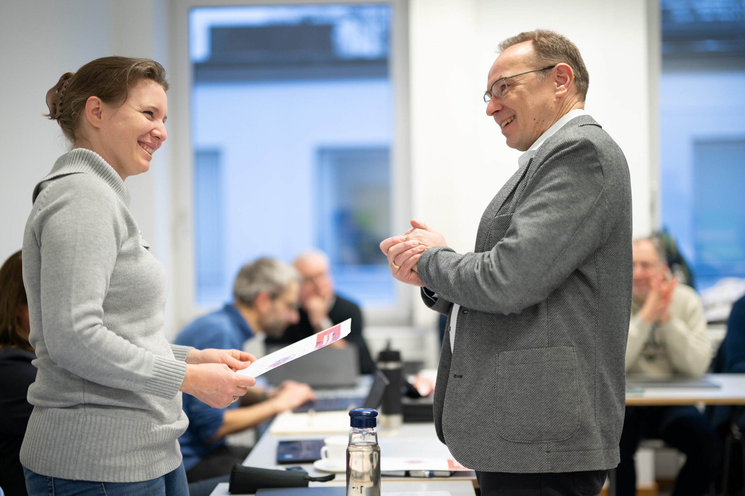 Two people are engaged in a conversation at Hochschule Coburg's office. The woman holds a piece of paper while the man smiles. In the background, other individuals are seated at tables, working or discussing.