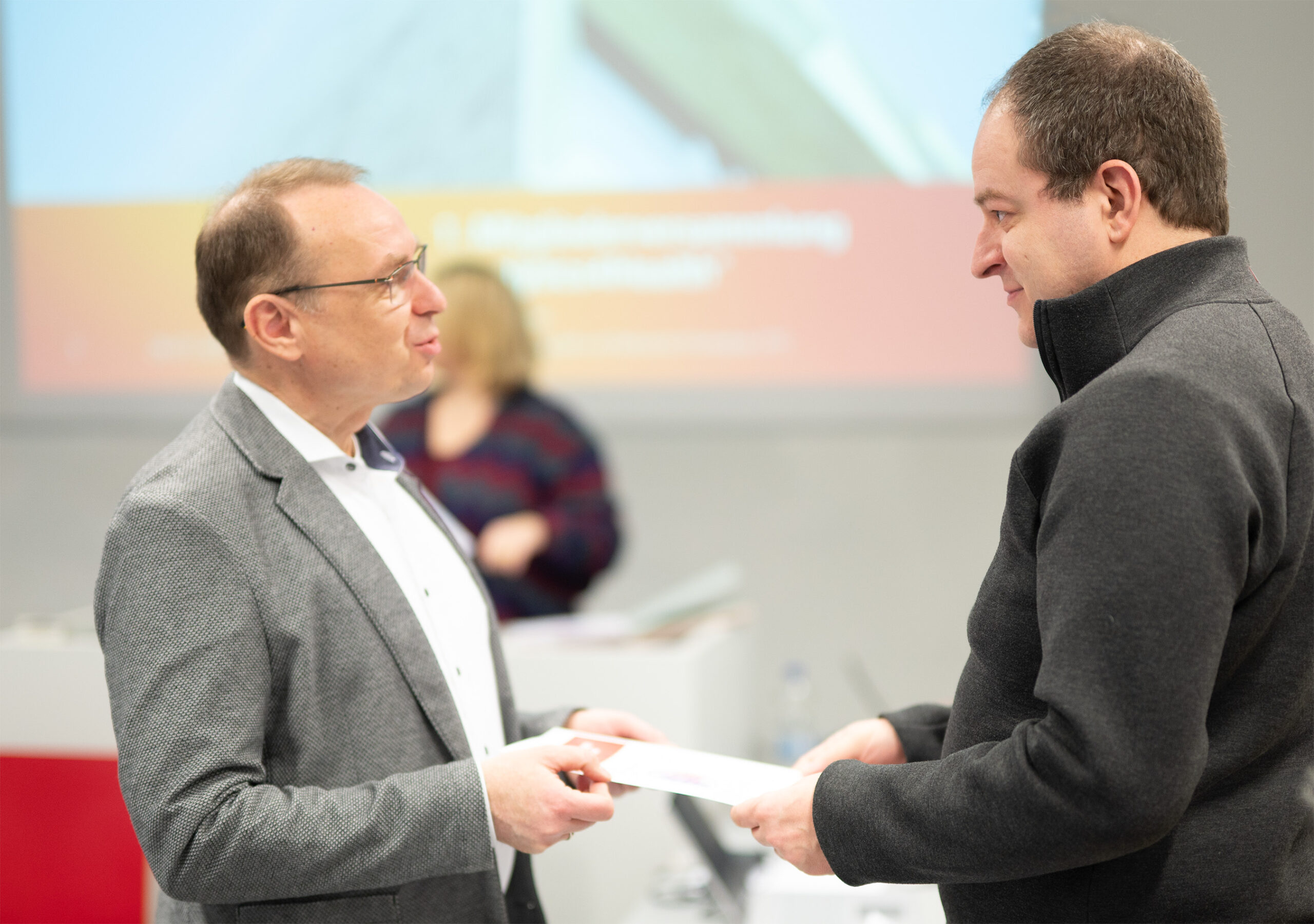 Two men are engaged in a conversation indoors at Hochschule Coburg. One holds a piece of paper, and a presentation screen is visible in the blurred background. Both men are dressed in business attire, reflecting the formal academic setting.