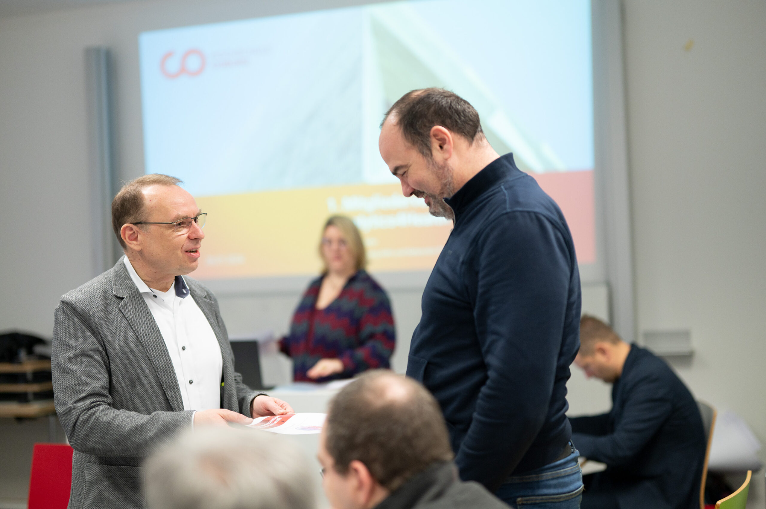 Two men from Hochschule Coburg are smiling and talking at an indoor event. One man is holding a piece of paper, while a woman stands in the background near a large presentation screen. Other attendees are seated nearby, engaged in the conference atmosphere.