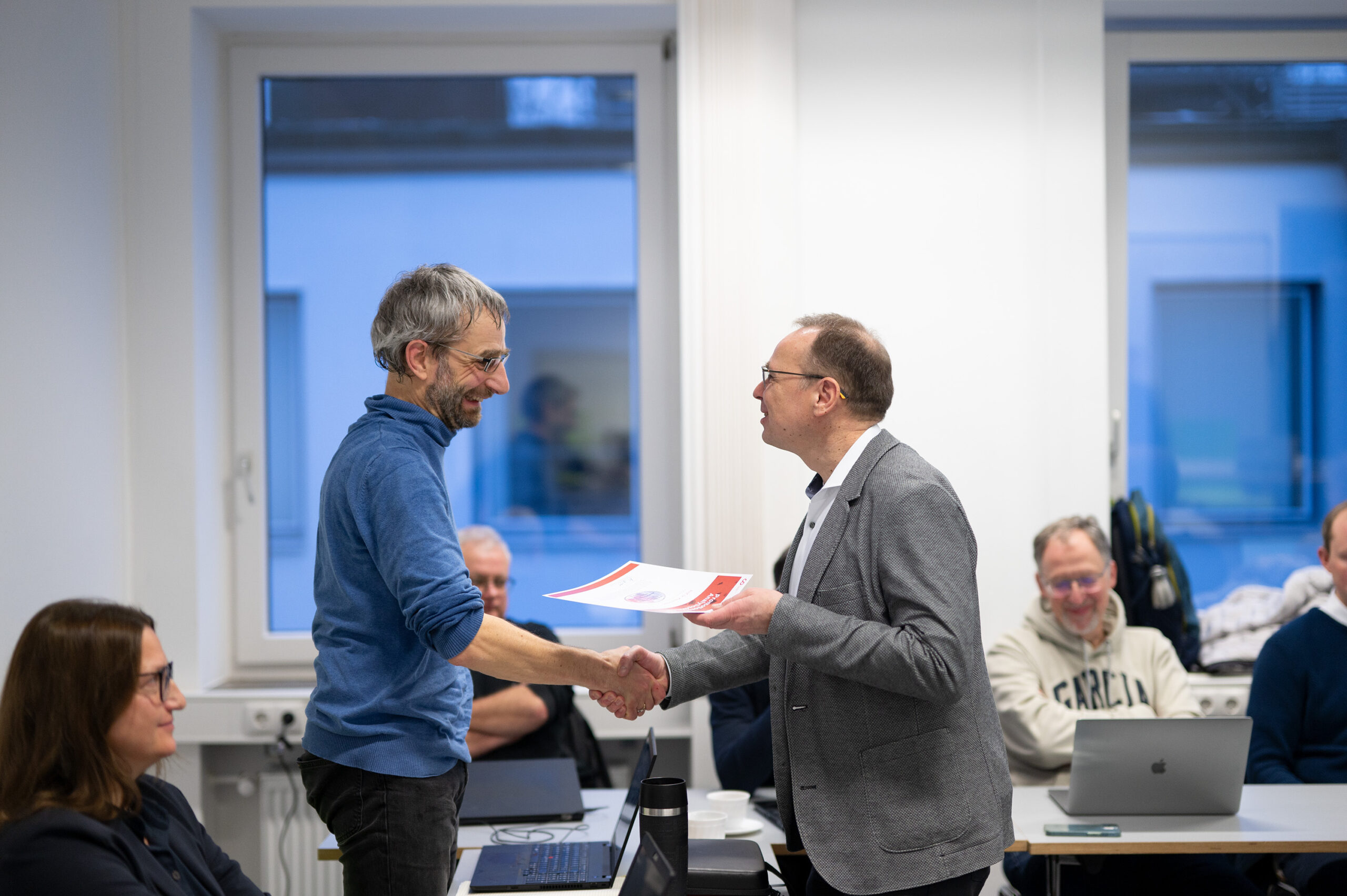 Two men are shaking hands in an office setting at Hochschule Coburg. One man is holding a certificate, while others are seated and observing the moment. A large window offers a glimpse of buildings outside, adding to the professional atmosphere.