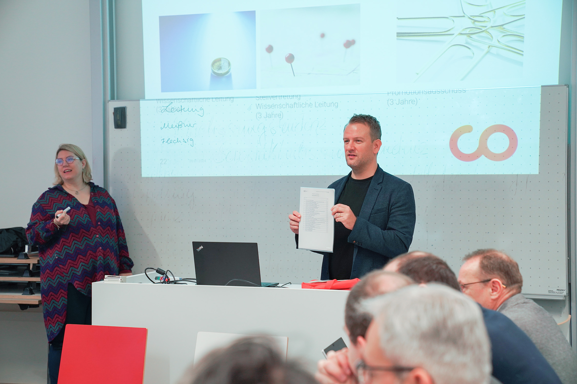 A man stands at a podium holding a sheet of paper while addressing an audience. A woman nearby smiles, and a presentation is displayed on a screen behind them, showcasing Hochschule Coburg. The room contains seated people.