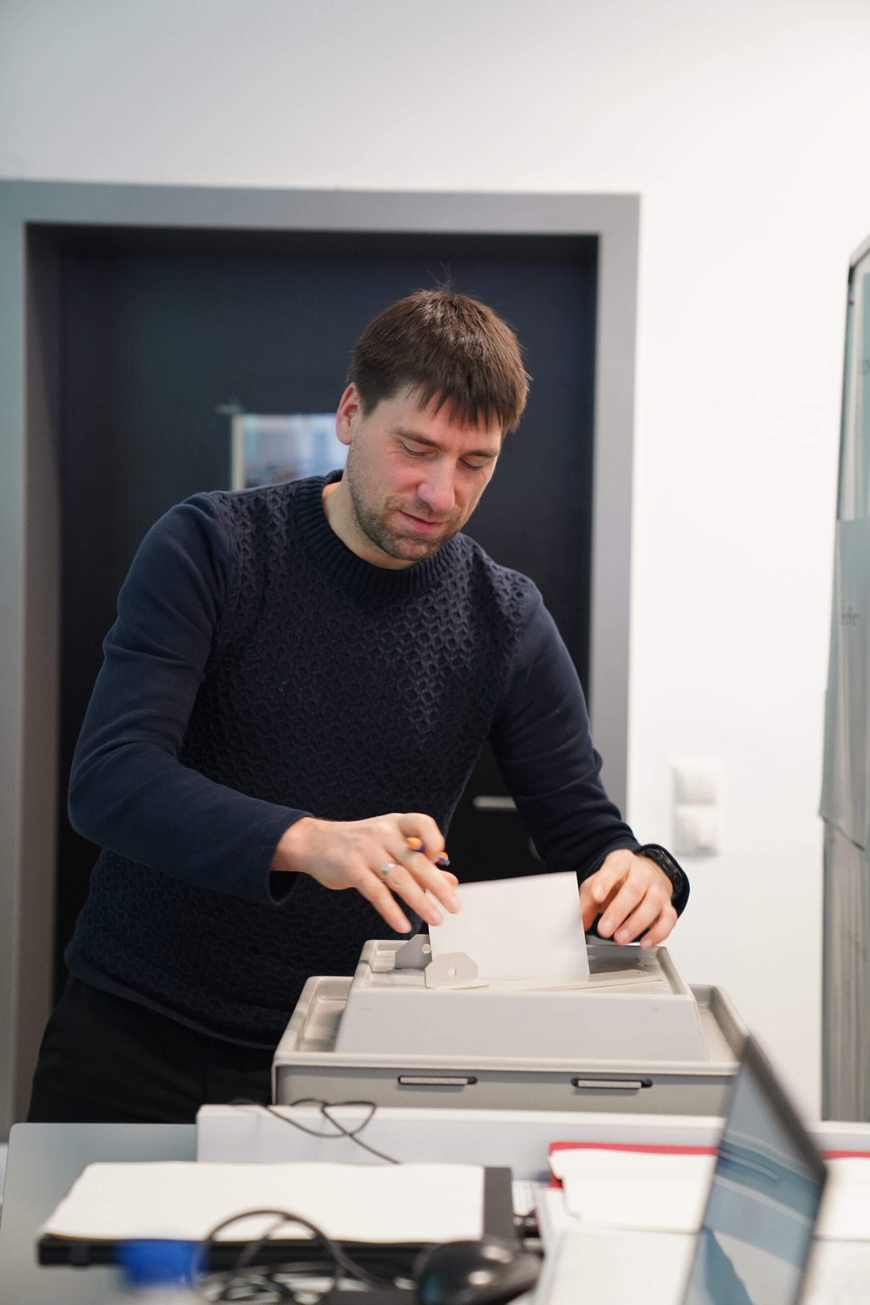 Inside a room at Hochschule Coburg, a person with short hair and clad in a dark sweater is placing a paper into a ballot box. The indoor setting reveals a door and some furniture in the background, capturing the essence of this academic environment.