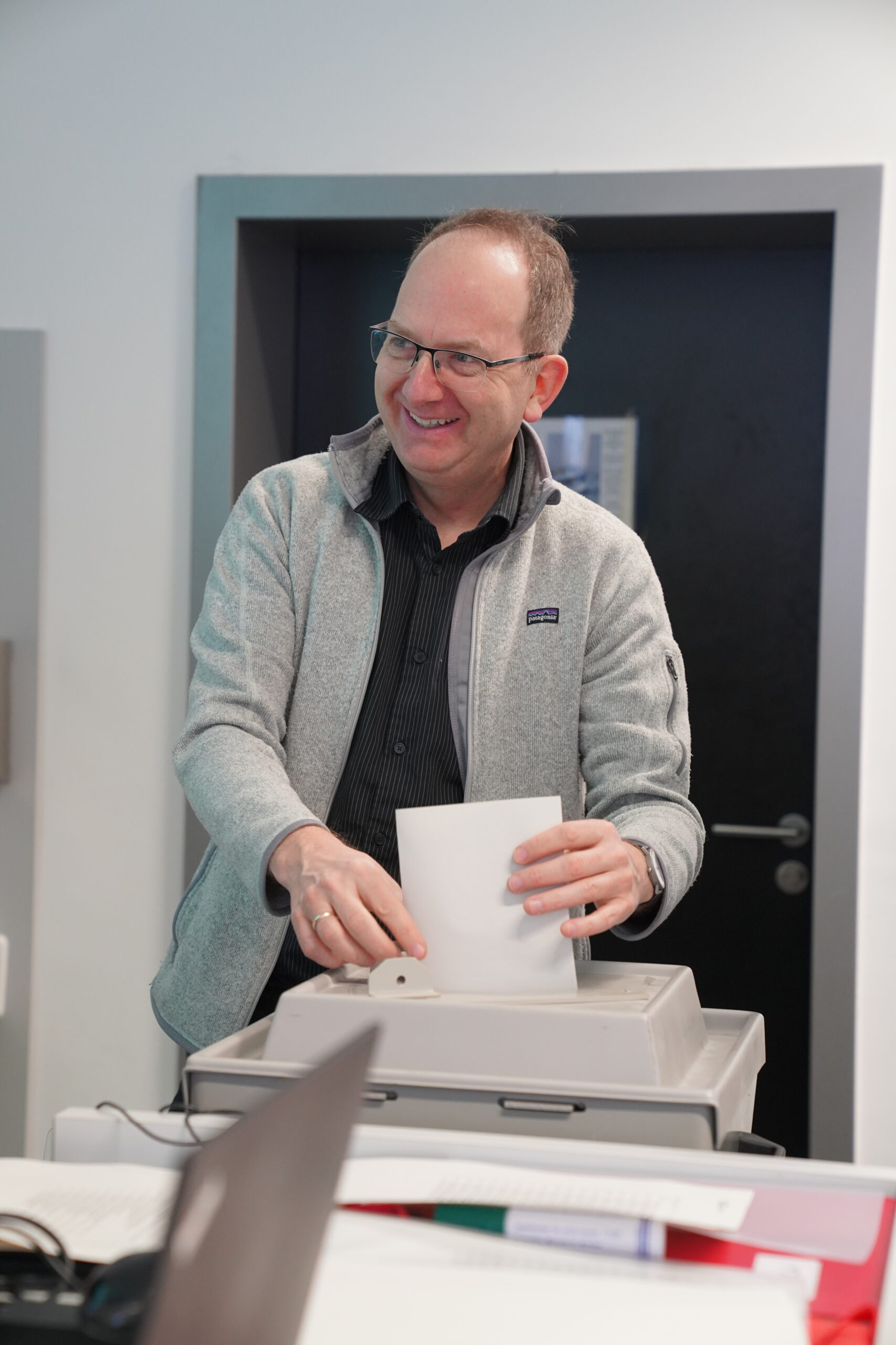 A person wearing glasses and a light gray jacket is smiling while inserting a piece of paper into a machine resembling a vote counting device at Hochschule Coburg. They stand in front of a dark door in the modern interior setting, embodying the spirit of innovation and academia.