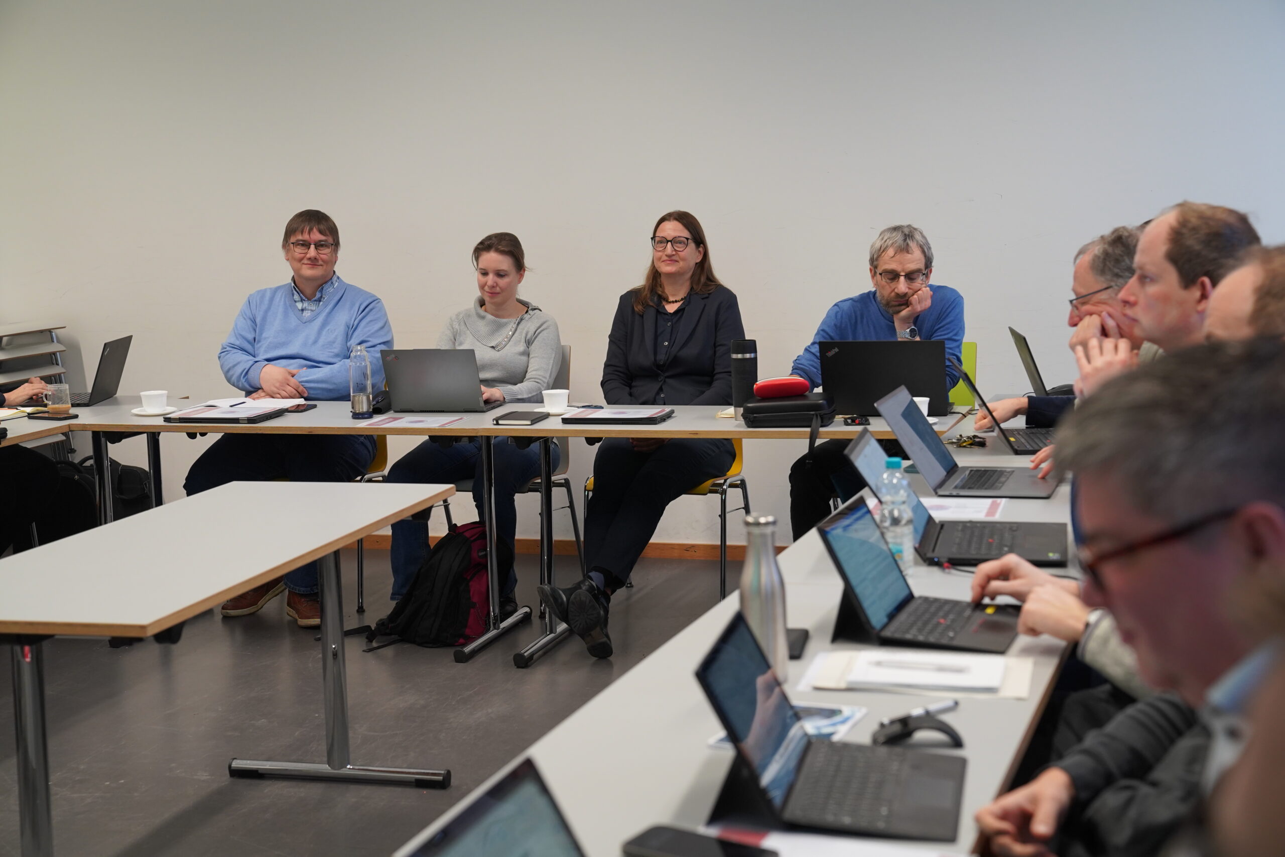 A group of people, possibly from Hochschule Coburg, sit around a conference table with laptops. Engaged in a lively discussion, they are surrounded by plain white walls with cups scattered across the table.