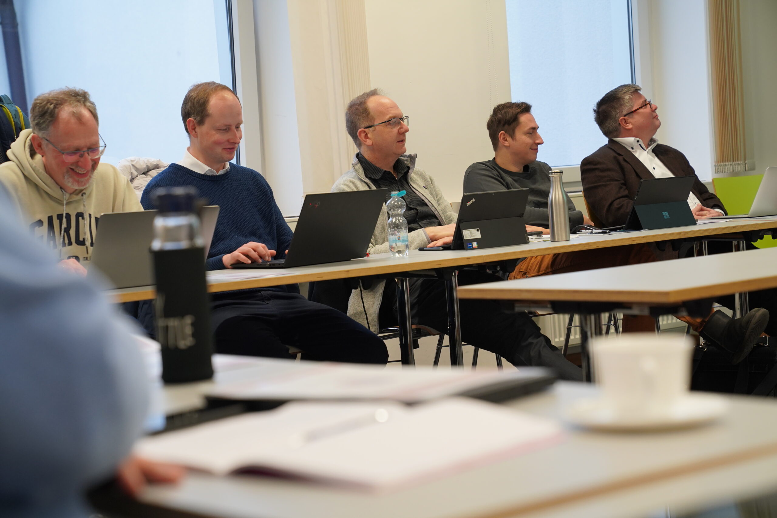 Five men sit at a long table in a conference room at Hochschule Coburg, each with a laptop in front of them. They appear engaged in a meeting or workshop, focused on their screens. A water bottle, cup, and papers are scattered across the table.