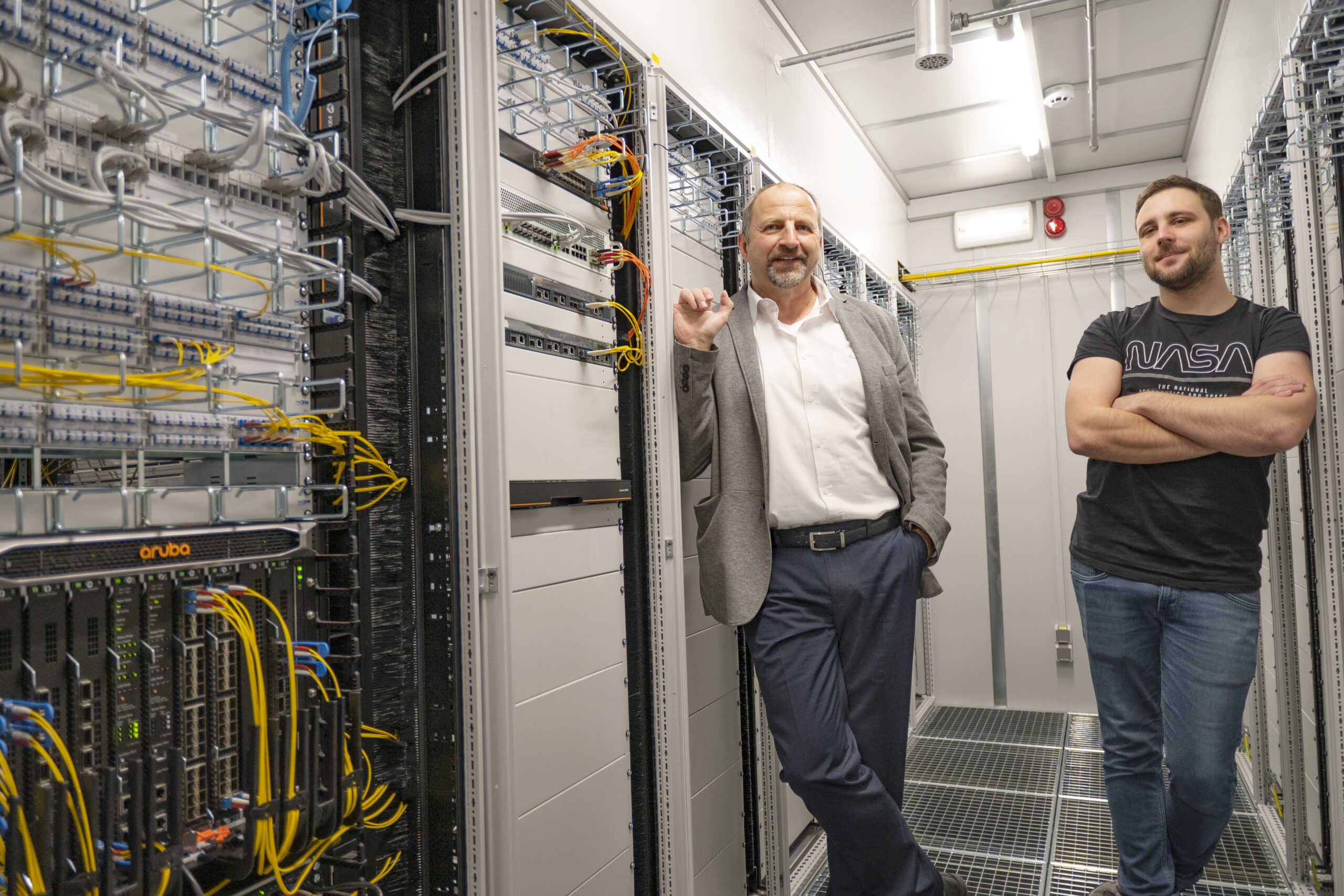 Zwei Männer stehen in einem Serverraum der Hochschule Coburg. Einer im Anzug lehnt an den Server-Racks, der andere steht im NASA-T-Shirt lässig mit verschränkten Armen inmitten eines Meeres aus Kabeln und Computerausrüstung.