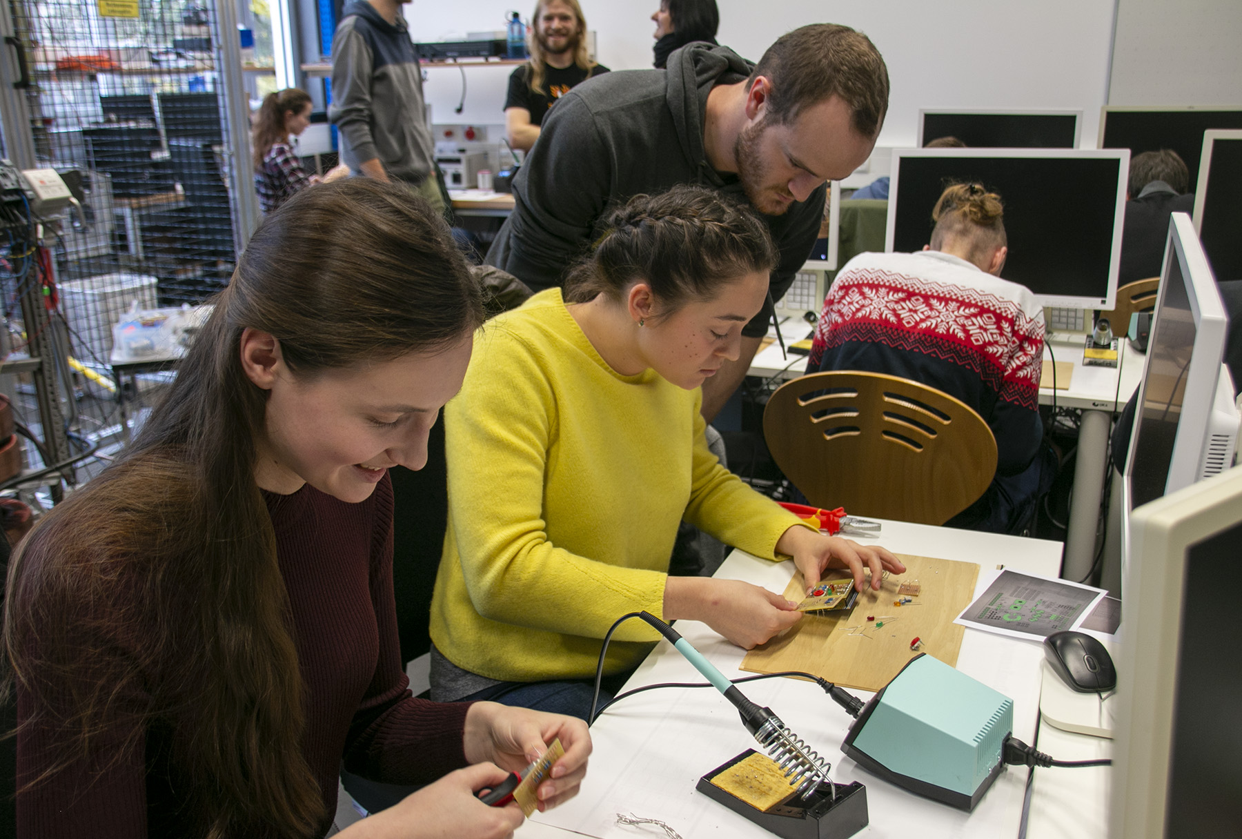 In einem Unterrichtsraum voller Lötgeräte und kleiner Platinen beschäftigt sich eine Gruppe intensiv mit einem Elektronik-Workshop an der Hochschule Coburg. Computer säumen die Tische, während andere Teilnehmer im Hintergrund konzentriert an ihren Aufgaben arbeiten, was zu einer lebendigen Lernatmosphäre beiträgt.