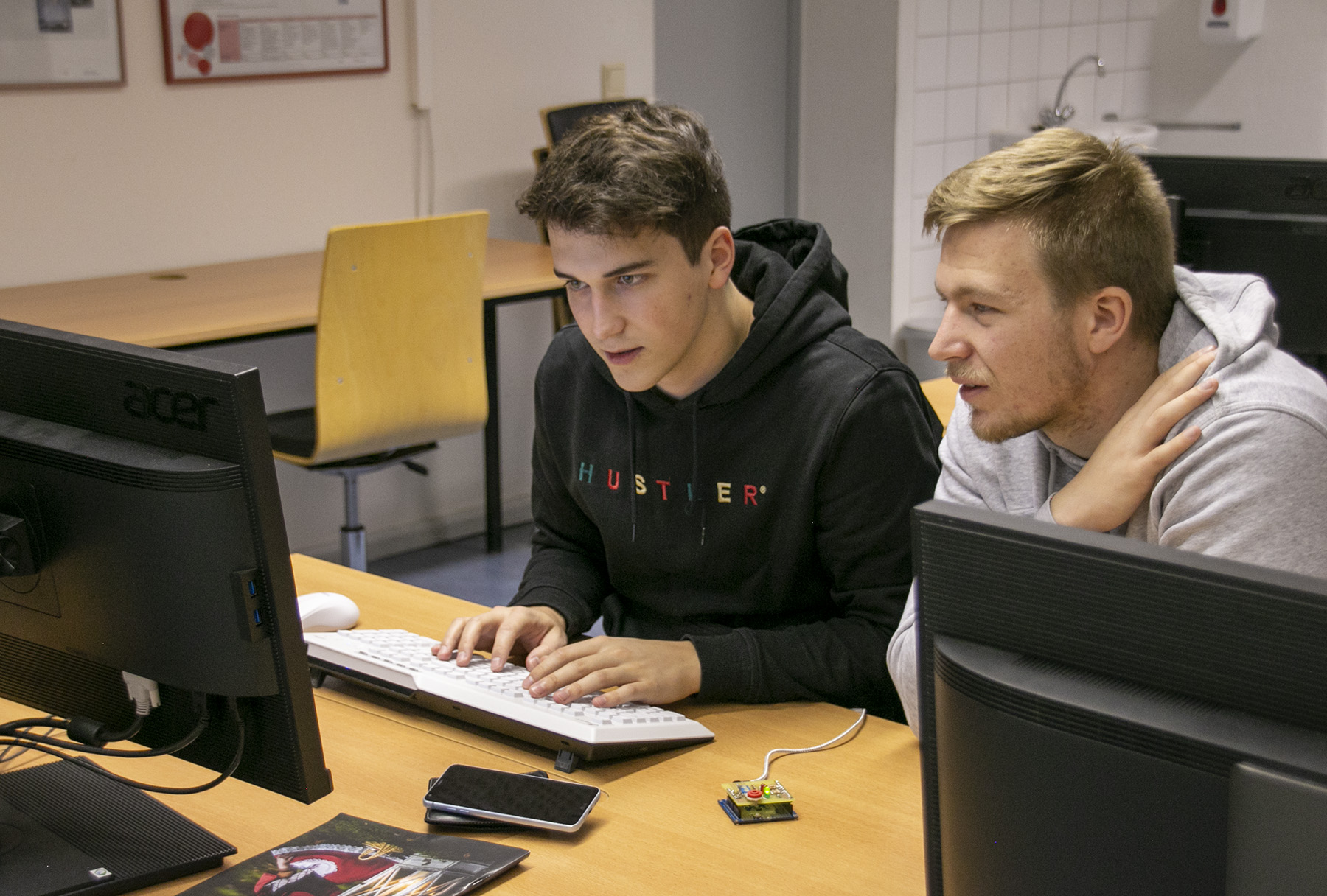 Zwei junge Männer der Hochschule Coburg sind in einen Computerbildschirm vertieft und tippen in einem Raum voller Schreibtische und einer weißen Tafel auf Tastaturen. Auf dem Schreibtisch neben ihnen liegt ein kleines elektronisches Bauteil, das auf ihr neuestes innovatives Projekt hindeutet.