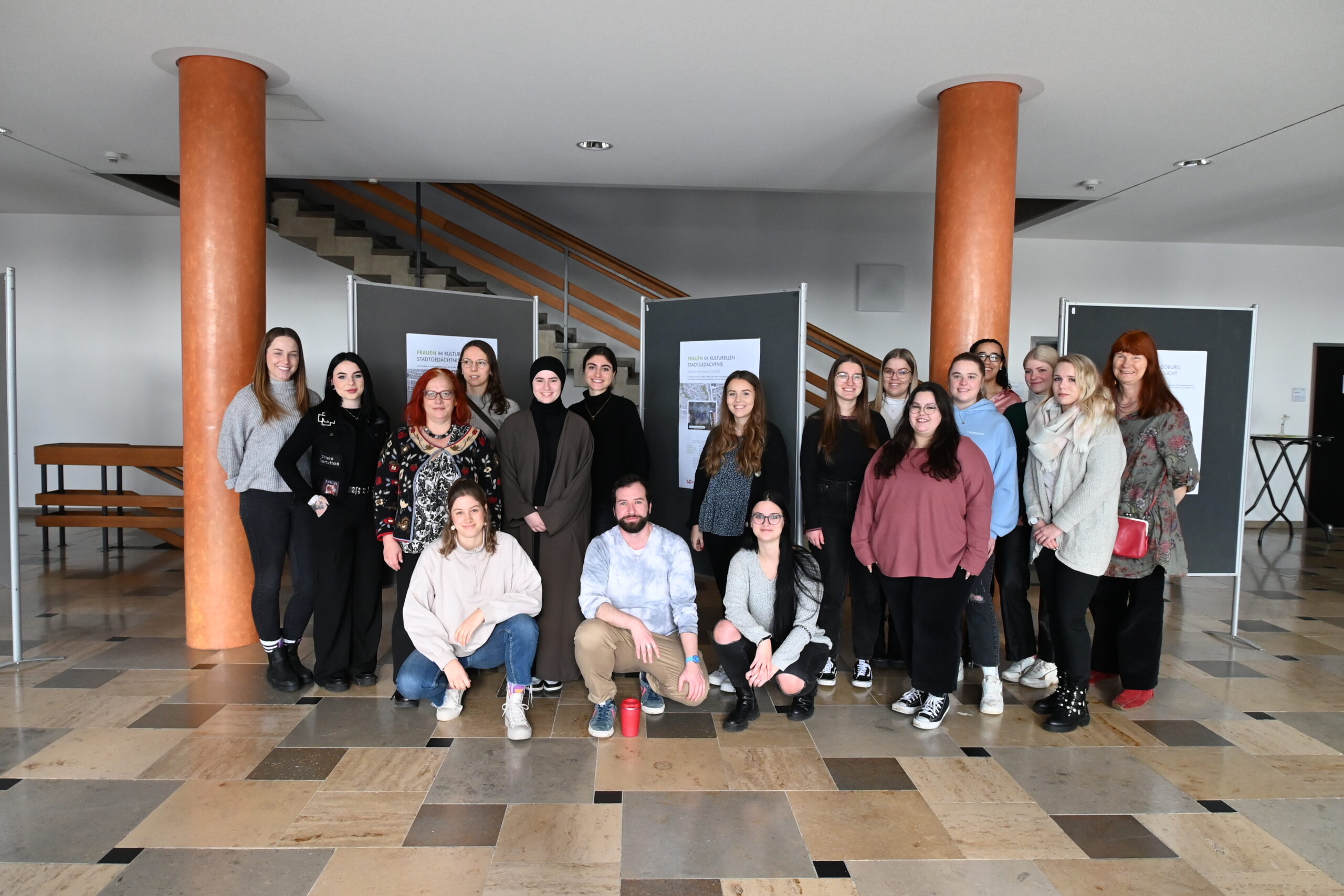 In einem großzügigen Raum mit Marmorboden und orangefarbenen Säulen posiert eine Gruppe von Studierenden der Hochschule Coburg vor Schautafeln für ein Foto. Einige stehen, einige knien in der ersten Reihe.