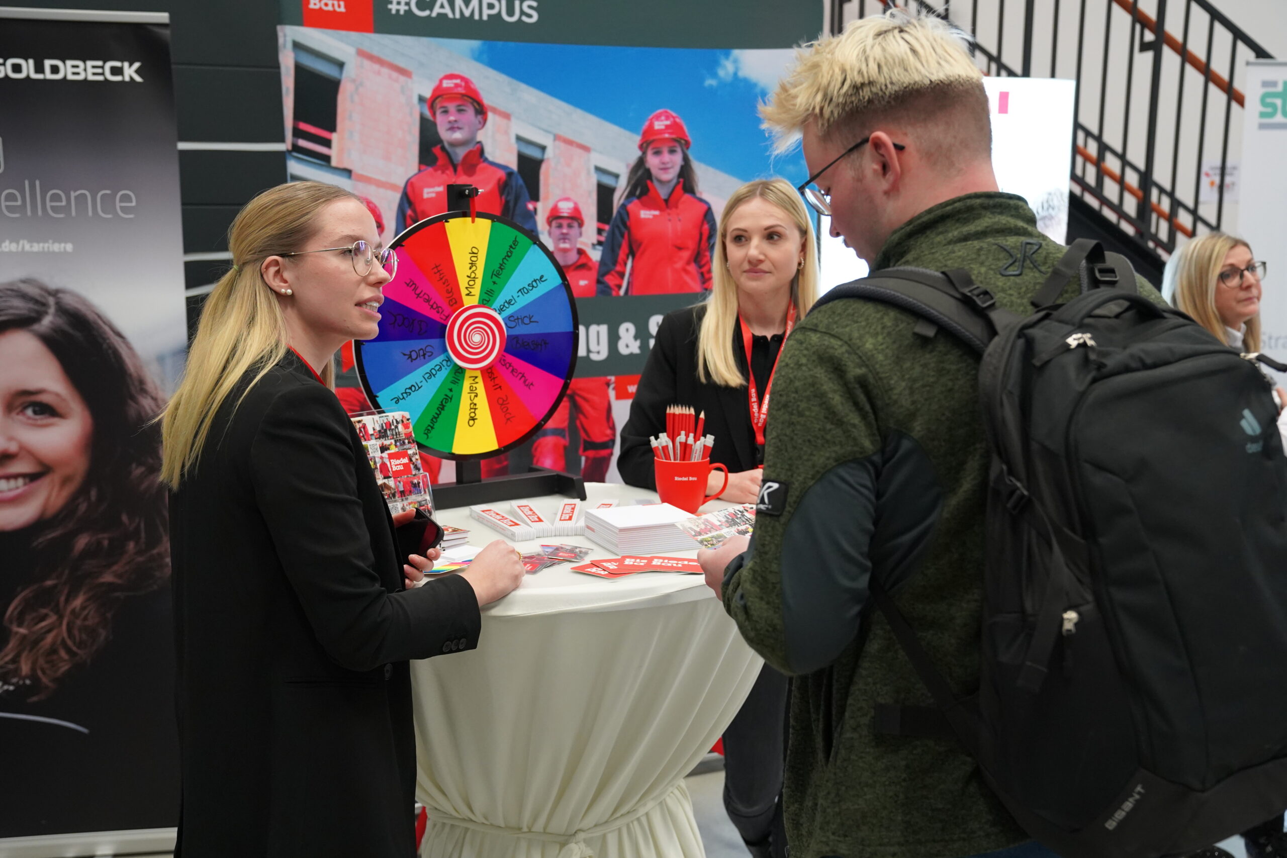 Menschen versammeln sich um einen Stand auf einer Messe oder Veranstaltung. Auf dem Tisch steht ein buntes Rad. Eine Frau spricht mit einem Mann, der einen Rucksack trägt, während andere zuschauen. Im Hintergrund sind Banner und Werbematerialien der Hochschule Coburg zu sehen, die der Szene einen akademischen Touch verleihen.