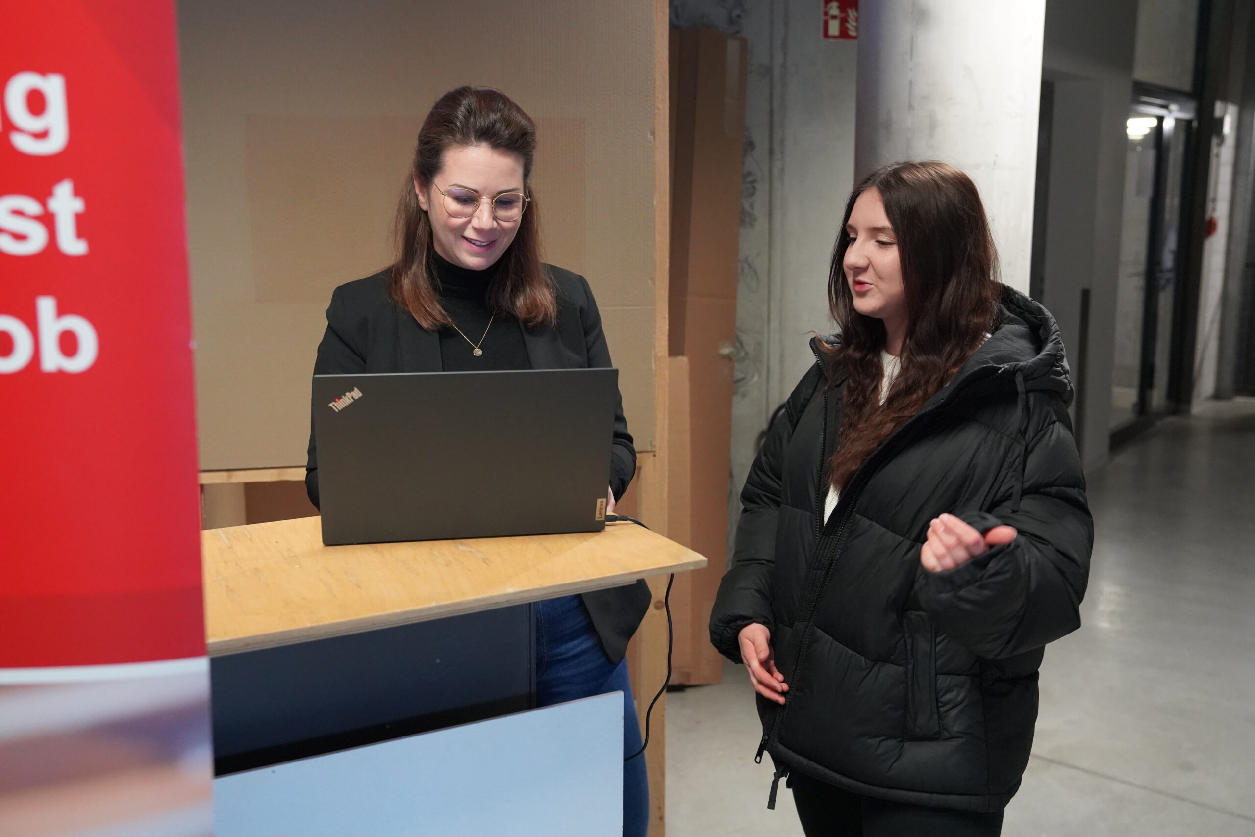 Two women are standing indoors at Hochschule Coburg. One is using a laptop on a wooden desk, smiling, while the other, wearing a black jacket, gestures with her hand. They appear to be engaged in a friendly conversation.