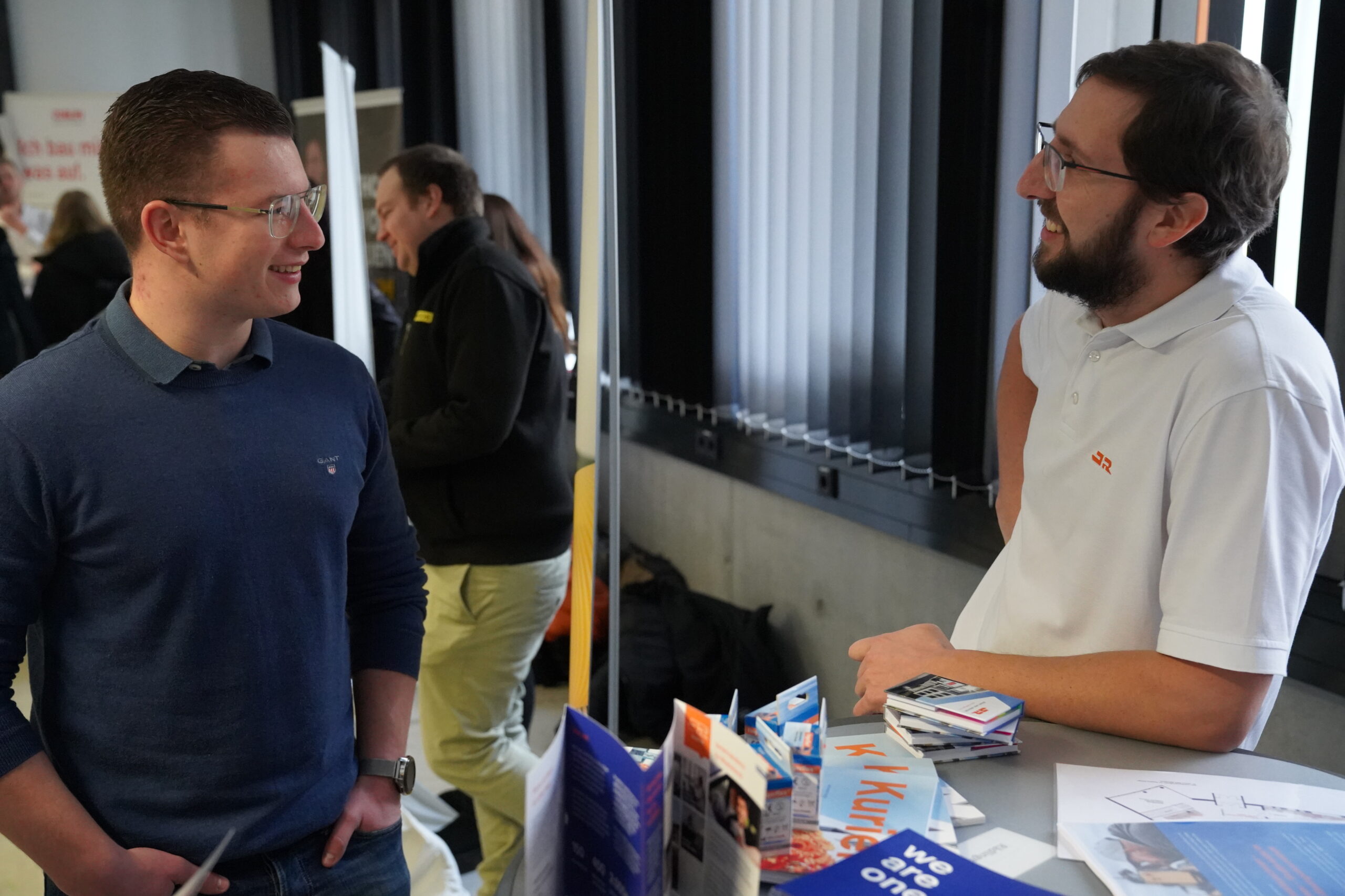 Two men are talking at a booth adorned with brochures and flyers, promoting Hochschule Coburg. One is wearing a blue sweater and glasses, the other a white polo shirt. They seem engaged in conversation, while a few other people and items are visible in the background.