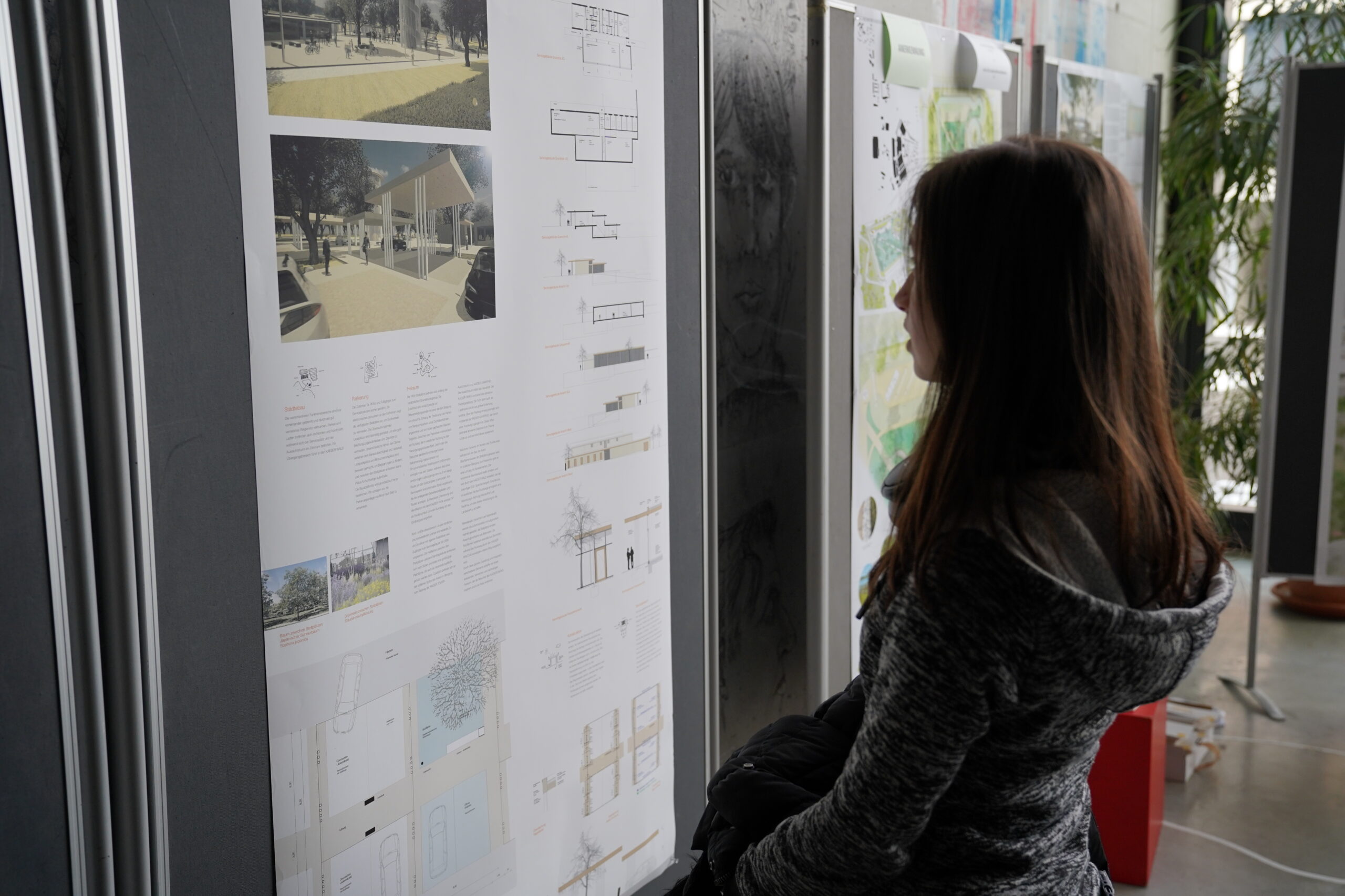 Eine Person mit langen Haaren und grauem Kapuzenpullover steht vor einer Schautafel in der Hochschule Coburg und betrachtet in einem gut beleuchteten Raum Architekturpläne und Diagramme.