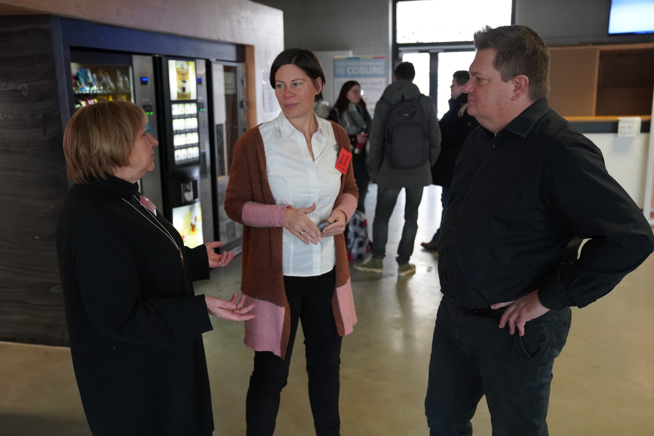 Three people are having a conversation indoors near vending machines at Hochschule Coburg. One woman, wearing a long cardigan, gestures as she speaks with a man in dark clothing and another woman in a coat. A few people are visible in the background.