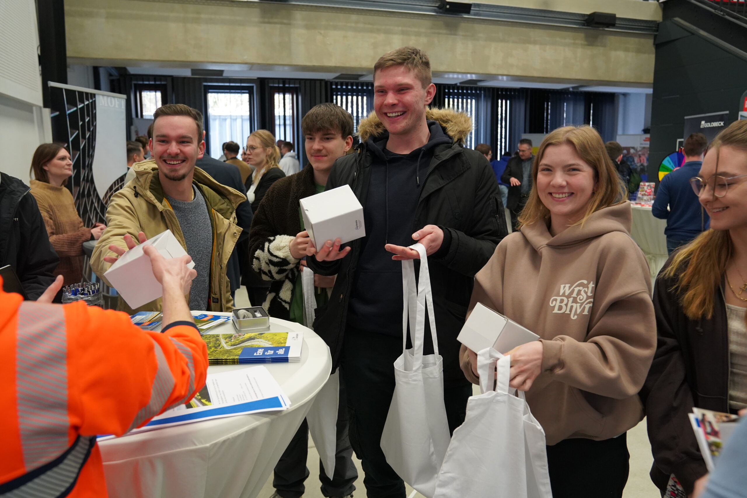 Bei einer Veranstaltung der Hochschule Coburg steht eine Gruppe lächelnder Menschen um einen Tisch und hält weiße Tragetaschen und Kartons in den Händen. Eine Person in einer orangefarbenen Jacke verteilt Geschenke. Im Hintergrund summt und strahlt die Atmosphäre der anderen Teilnehmer Begeisterung und Gemeinschaftsgeist aus.