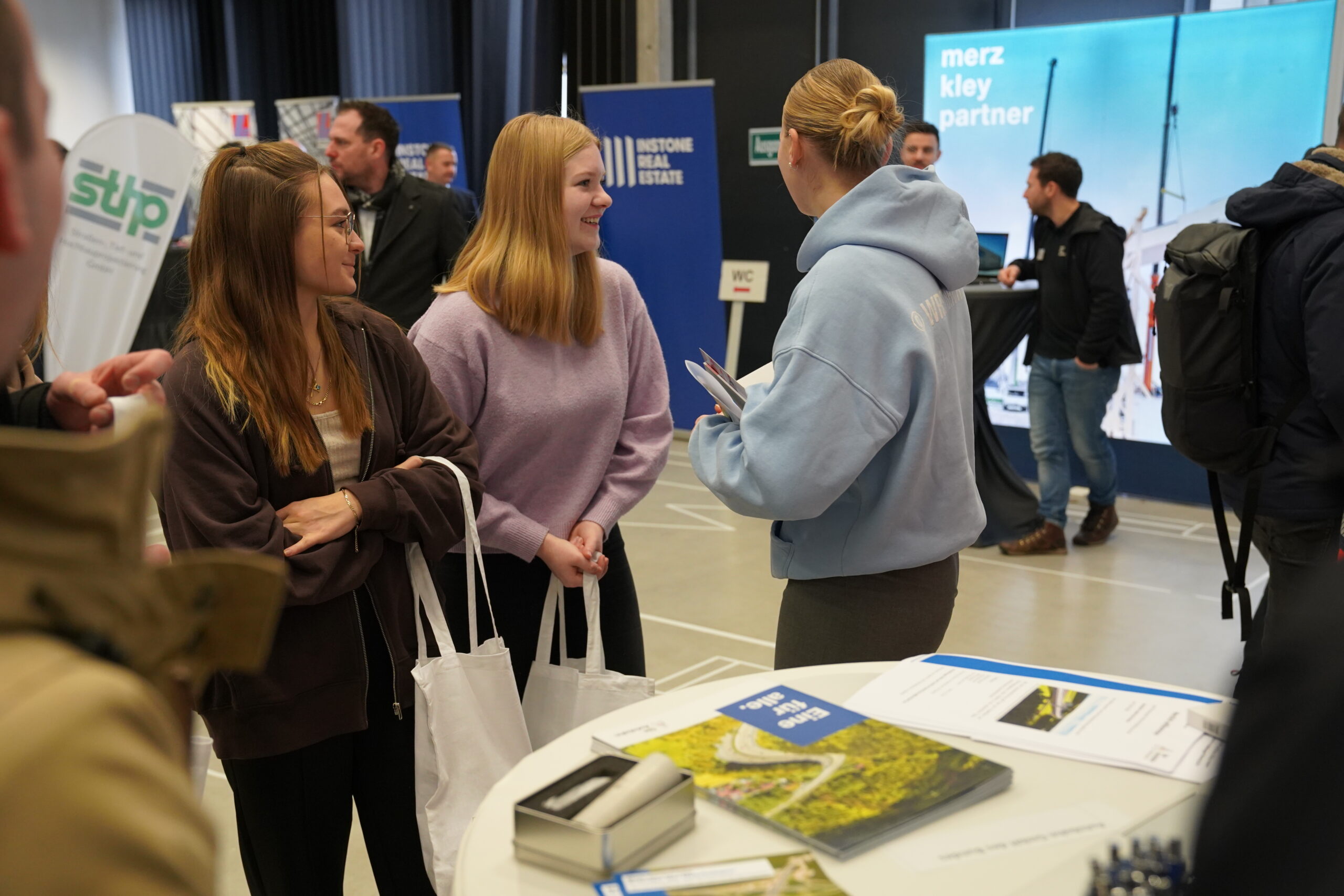 At a bustling career fair, a group engages earnestly, with two young women holding tote bags intently listening to another in a blue hoodie from Hochschule Coburg. Brochures scatter the nearby table as company banners form a vibrant backdrop.