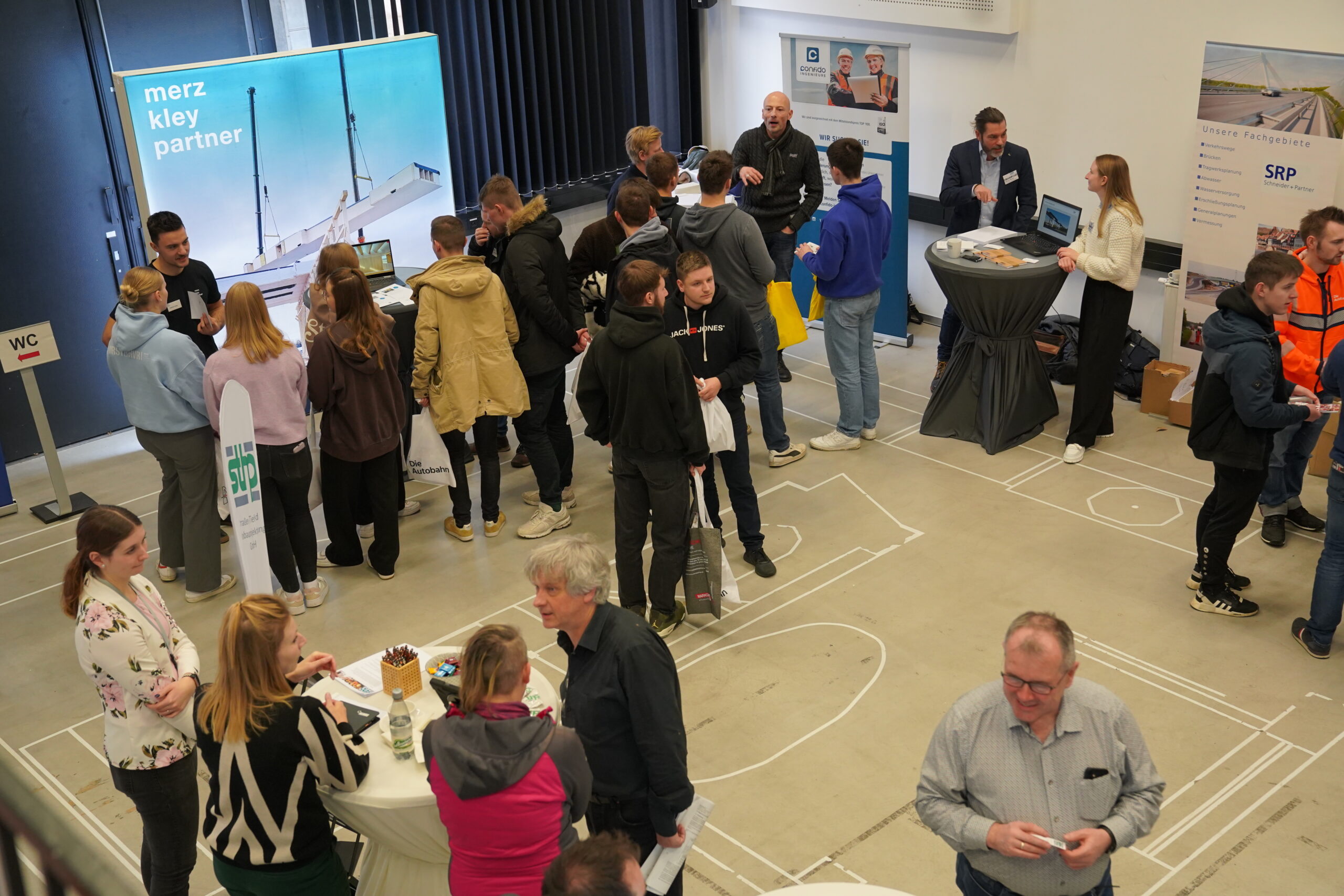 Bei einer geschäftigen Veranstaltung in Innenräumen der Hochschule Coburg diskutieren Gruppen. An mehreren Ständen sind Banner aufgestellt, an denen Einzelpersonen stehen oder sitzen, der Boden ist mit Linien markiert. Einige Teilnehmer halten Broschüren in den Händen, während auf den Laptops vor Aktivität gebrummt wird.