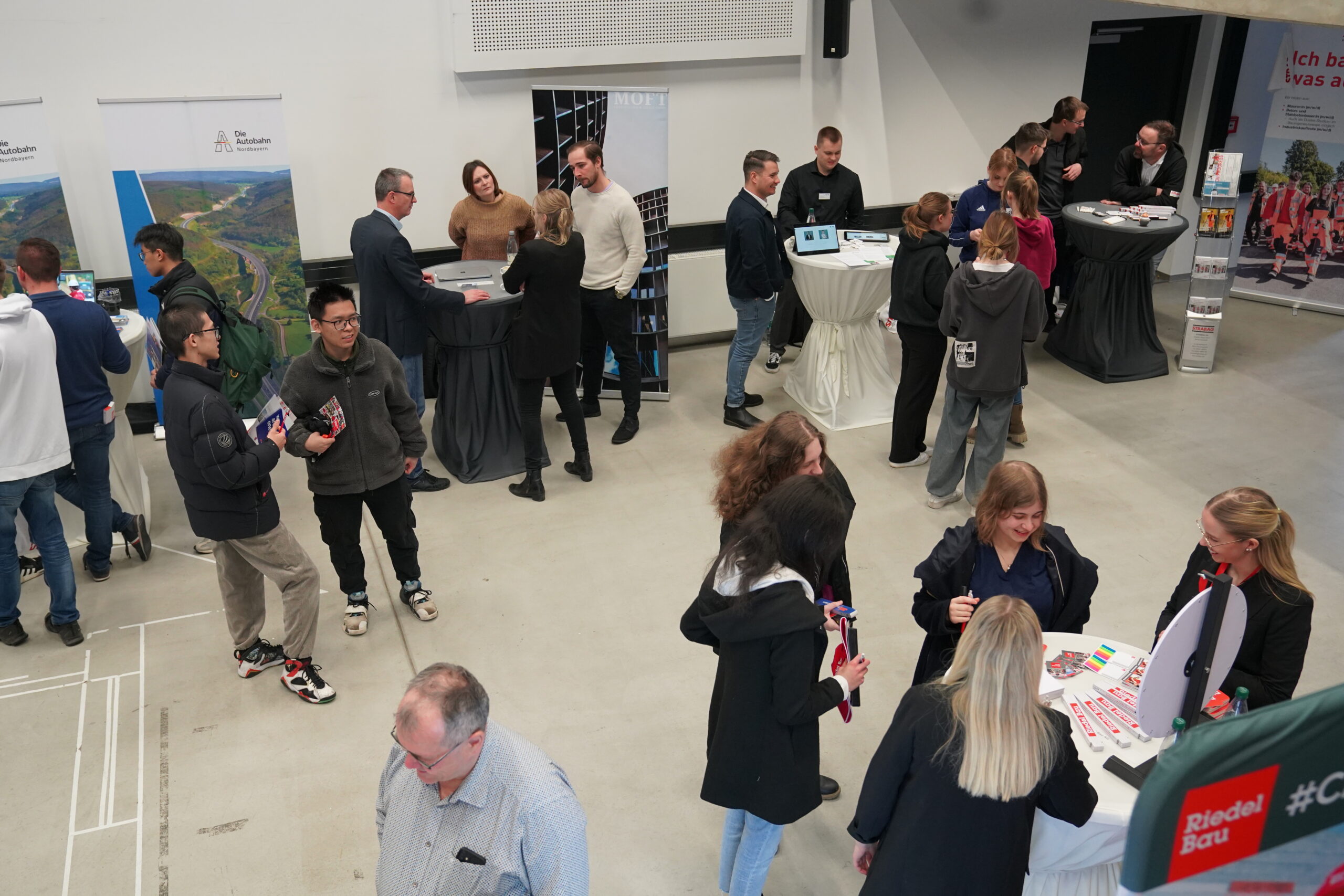 People are gathered in a room with various booths and tables at the Hochschule Coburg expo. Some are talking, while others examine pamphlets or products. The setting buzzes with networking energy, complete with banners and informational displays in the background.