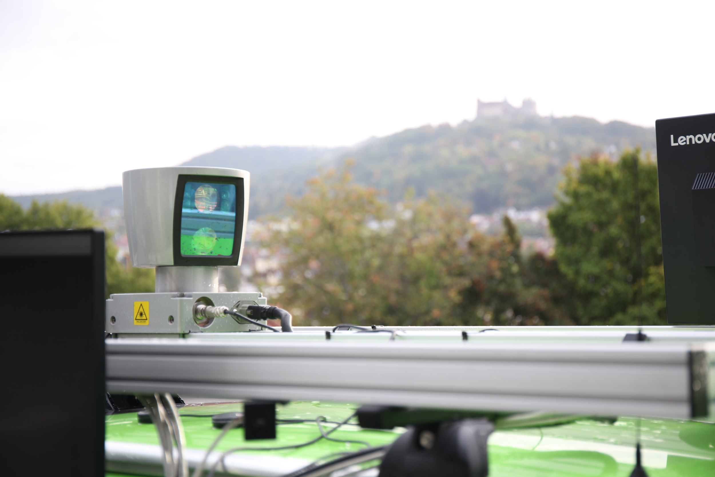 Ein grünes Fahrzeug, das oben mit einem Scanner mit dem Logo der Hochschule Coburg ausgestattet ist, verfügt über einen kleinen Bildschirm, auf dem Daten angezeigt werden. Im Hintergrund zeichnet sich unter einem wolkigen Himmel eine verschwommene Landschaft mit Hügeln und einem Gebäude ab.