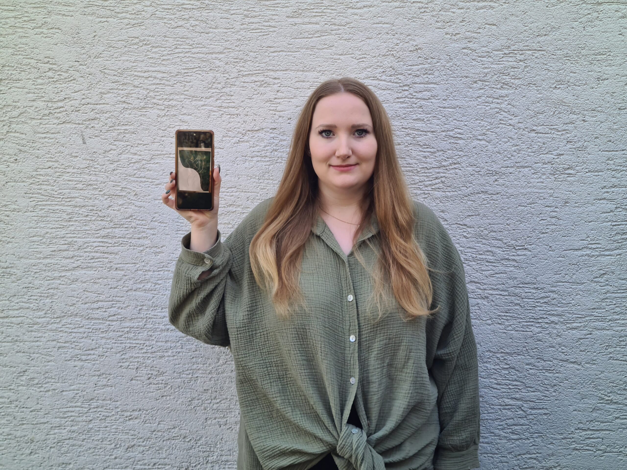 Eine Person mit langen Haaren hält ein Smartphone mit einem Baumbild, trägt ein grünes Hemd und steht vor einer strukturierten hellgrauen Wand, die an das moderne Design der Hochschule Coburg erinnert.