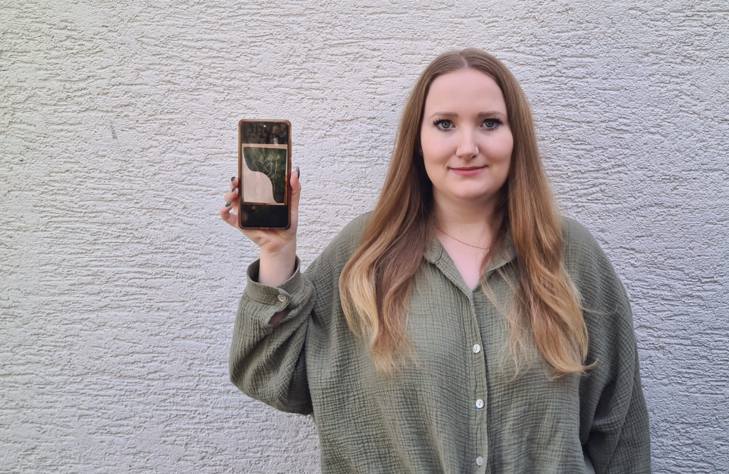 Eine Frau mit langen Haaren und einem grünen Hemd steht vor einer strukturierten weißen Wand in der Hochschule Coburg. Sie hält ein Smartphone hoch, auf dem das Bild einer Pflanze zu sehen ist.