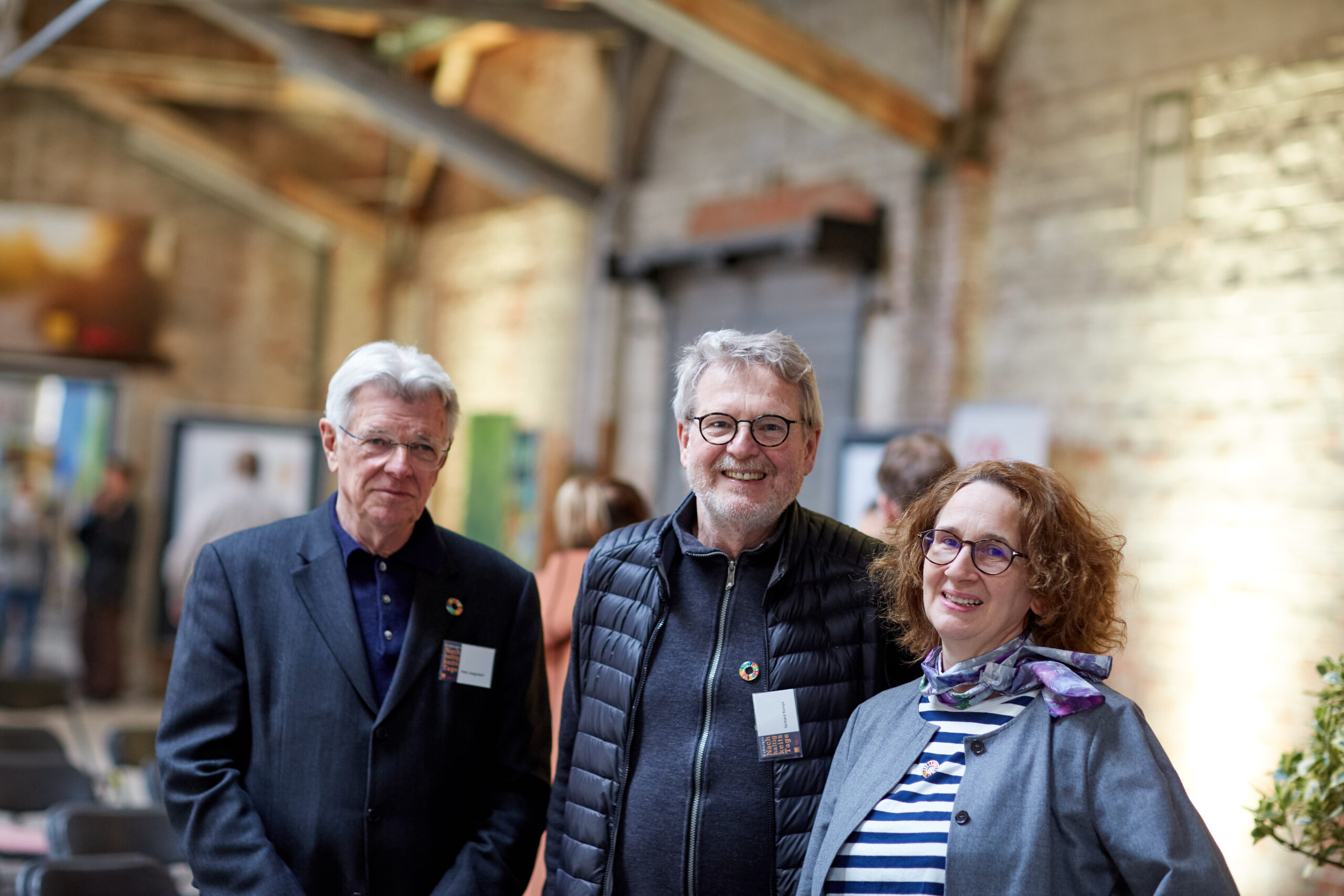 Three people with Hochschule Coburg name tags are smiling and standing together indoors at an event. The room's exposed brick walls and beams create a rustic atmosphere, complementing their casual business attire.