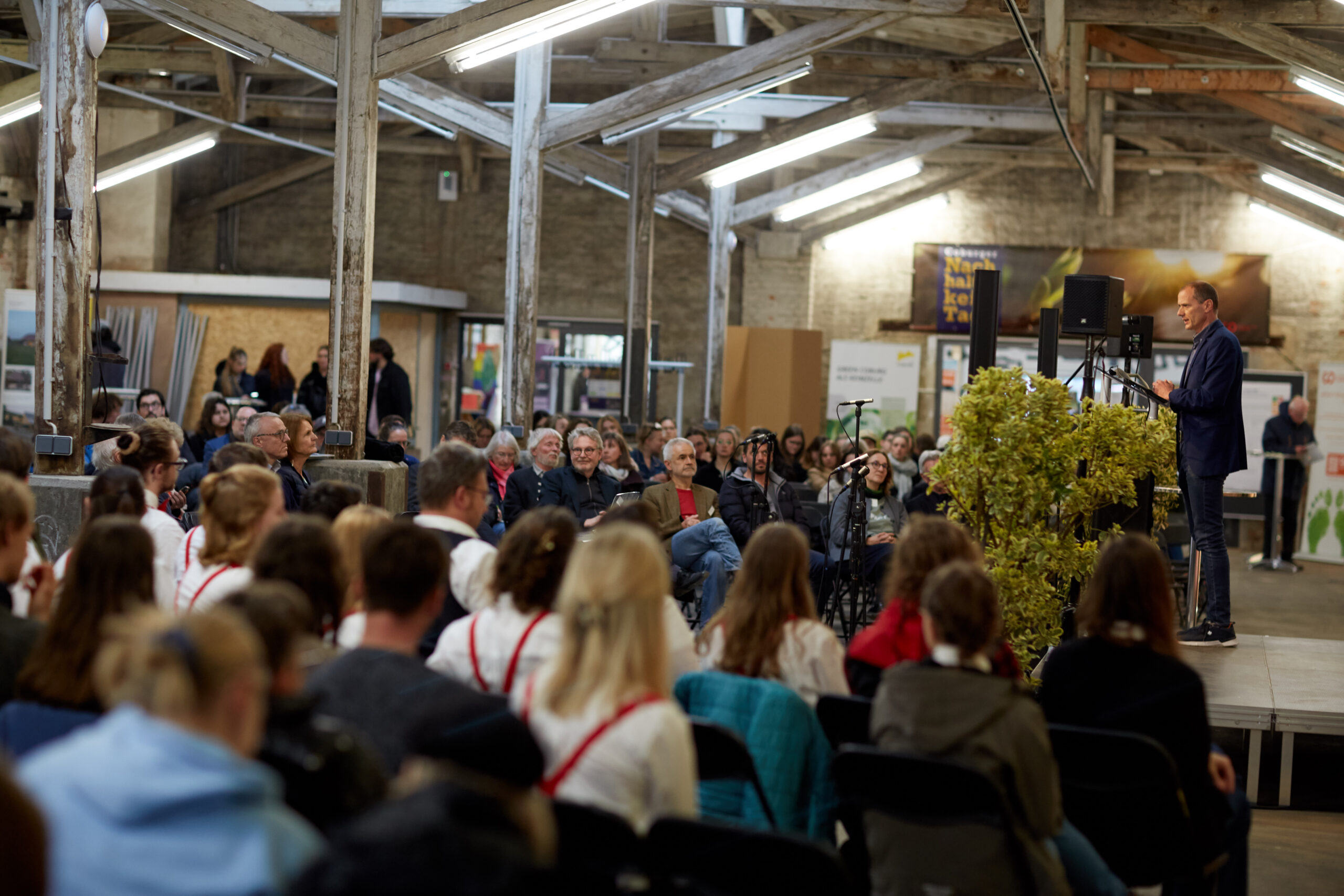 A speaker at Hochschule Coburg addresses a large audience seated in an industrial-style room with exposed beams. The crowd listens attentively, some taking notes. A podium adorned with greenery stands amidst natural light streaming through the windows.