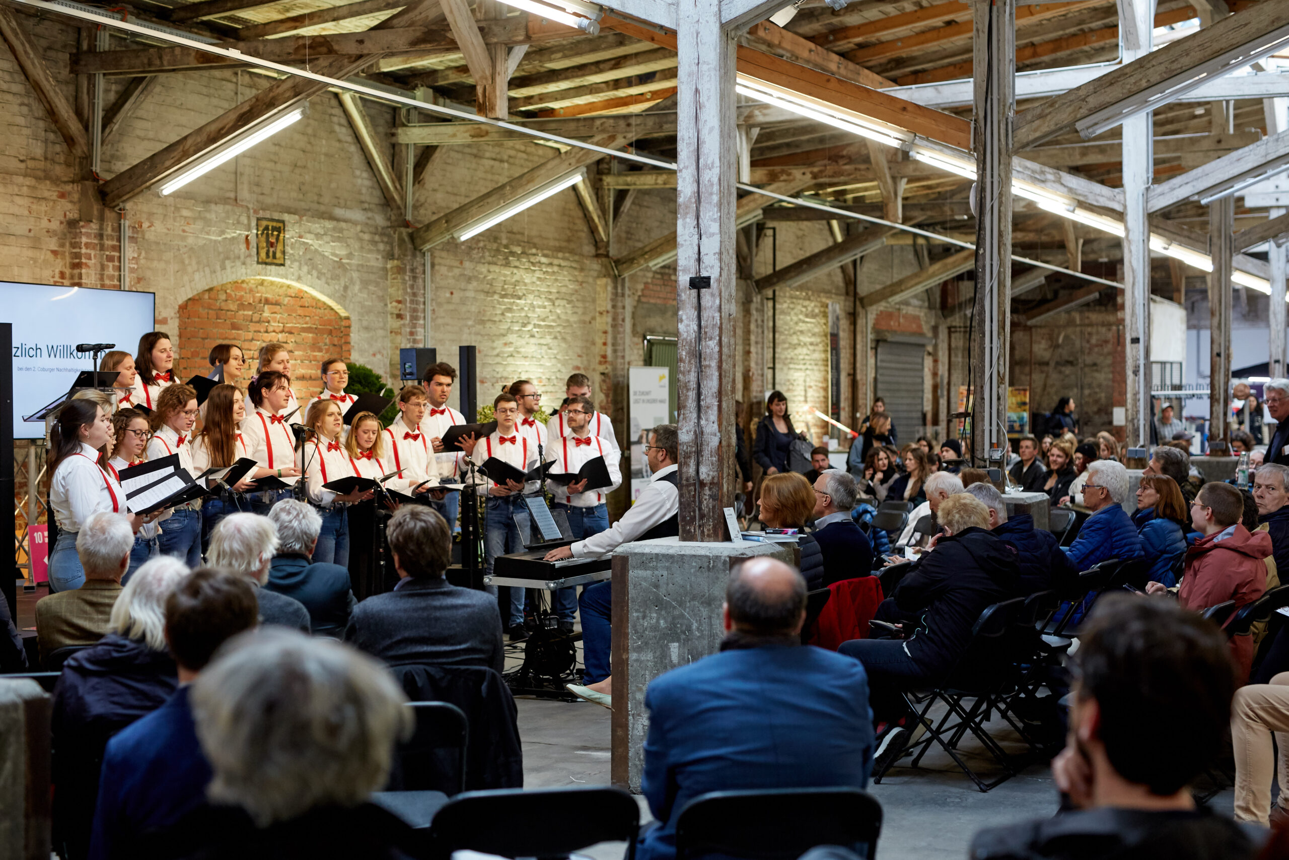 In a rustic industrial venue with wooden beams and brick walls, the Hochschule Coburg choir performs beautifully. The conductor guides them as an audience watches attentively, with choir members donning black and white outfits accented by red bow ties.