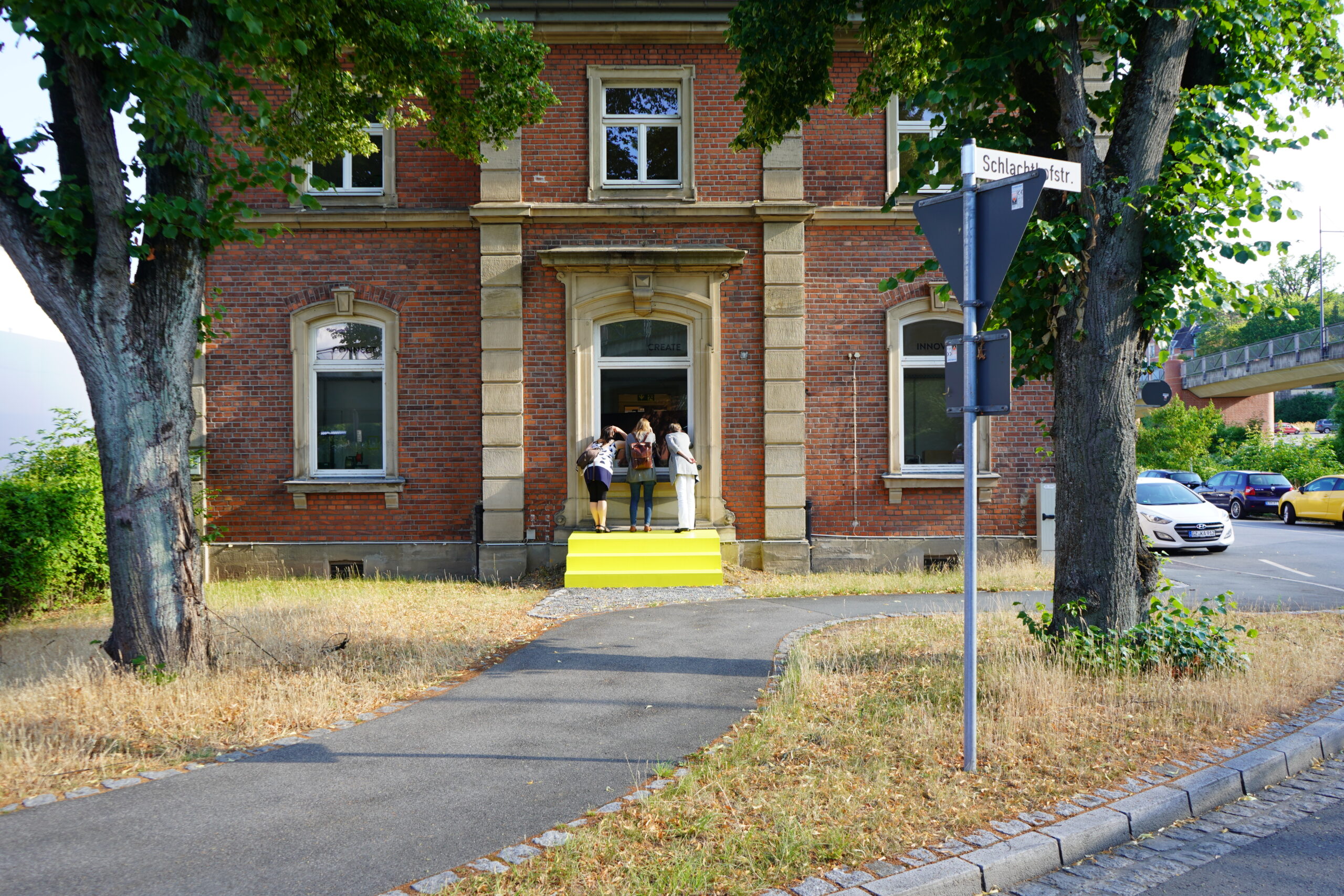 Eine Gruppe von Menschen steht auf den Stufen eines Backsteingebäudes mit Bogenfenstern und einem gelb gestrichenen Eingang, der an den Campus der Hochschule Coburg erinnert. Das Gebäude ist von Bäumen und Gras eingerahmt, in der Nähe gibt es einen Bürgersteig und ein paar geparkte Autos.