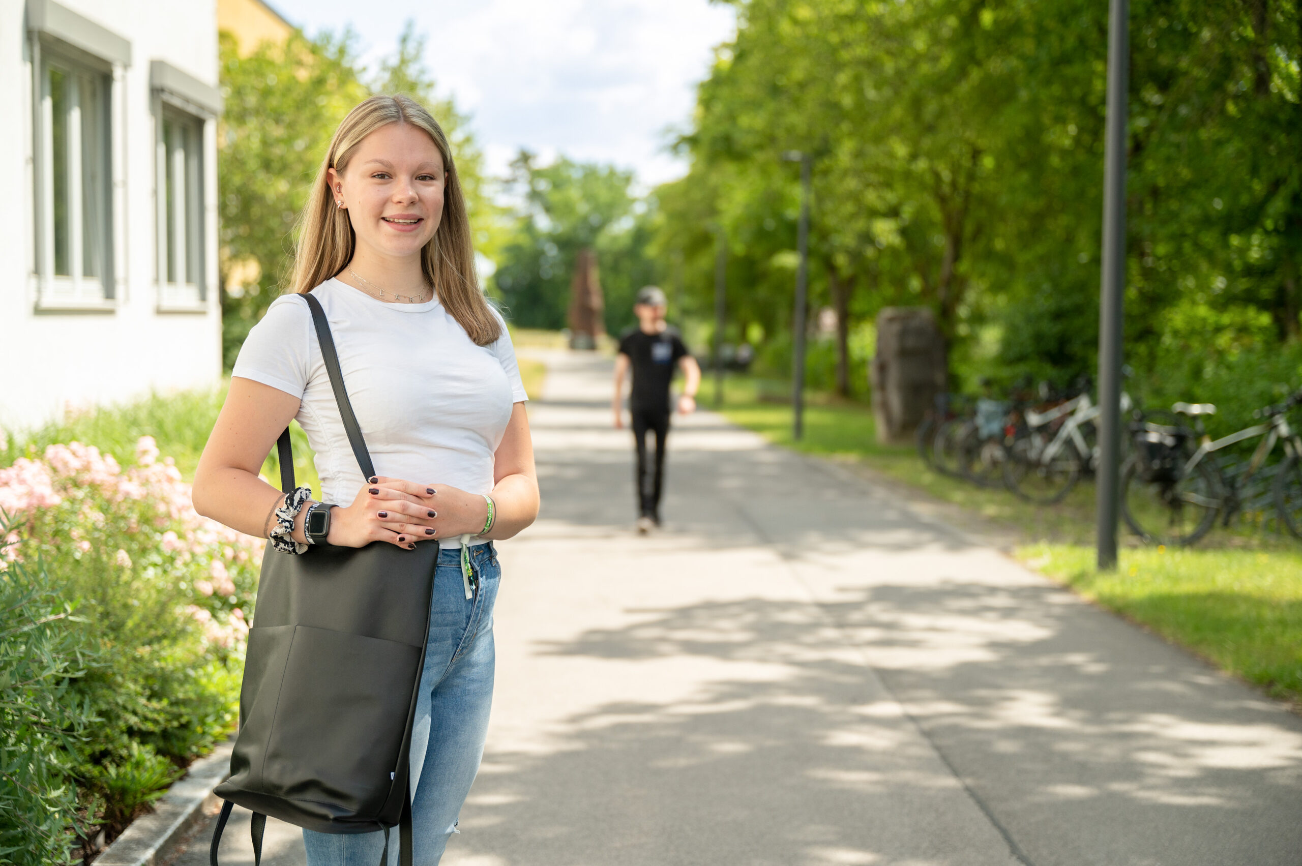 Eine Person mit langen Haaren, bekleidet mit einem weißen Hemd und Jeans, steht auf einem sonnigen Weg in der Nähe der Hochschule Coburg und hält eine schwarze Tasche in der Hand. Im Hintergrund schlendert eine Gestalt in Schwarz spazieren, während Fahrräder entlang des von Grün gesäumten Weges stehen.